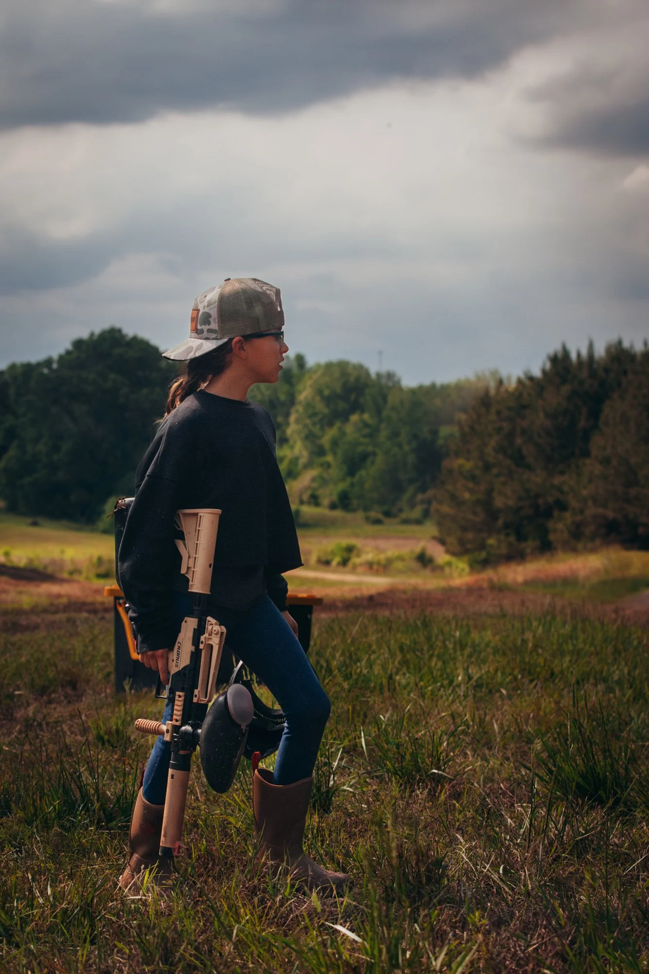A woman with glasses, wearing a cap, black sweater, blue jeans, and tall boots, standing in a field with a rifle in her hand and a firearm holstered on her thigh, overlooking a landscape with trees and cloudy sky.