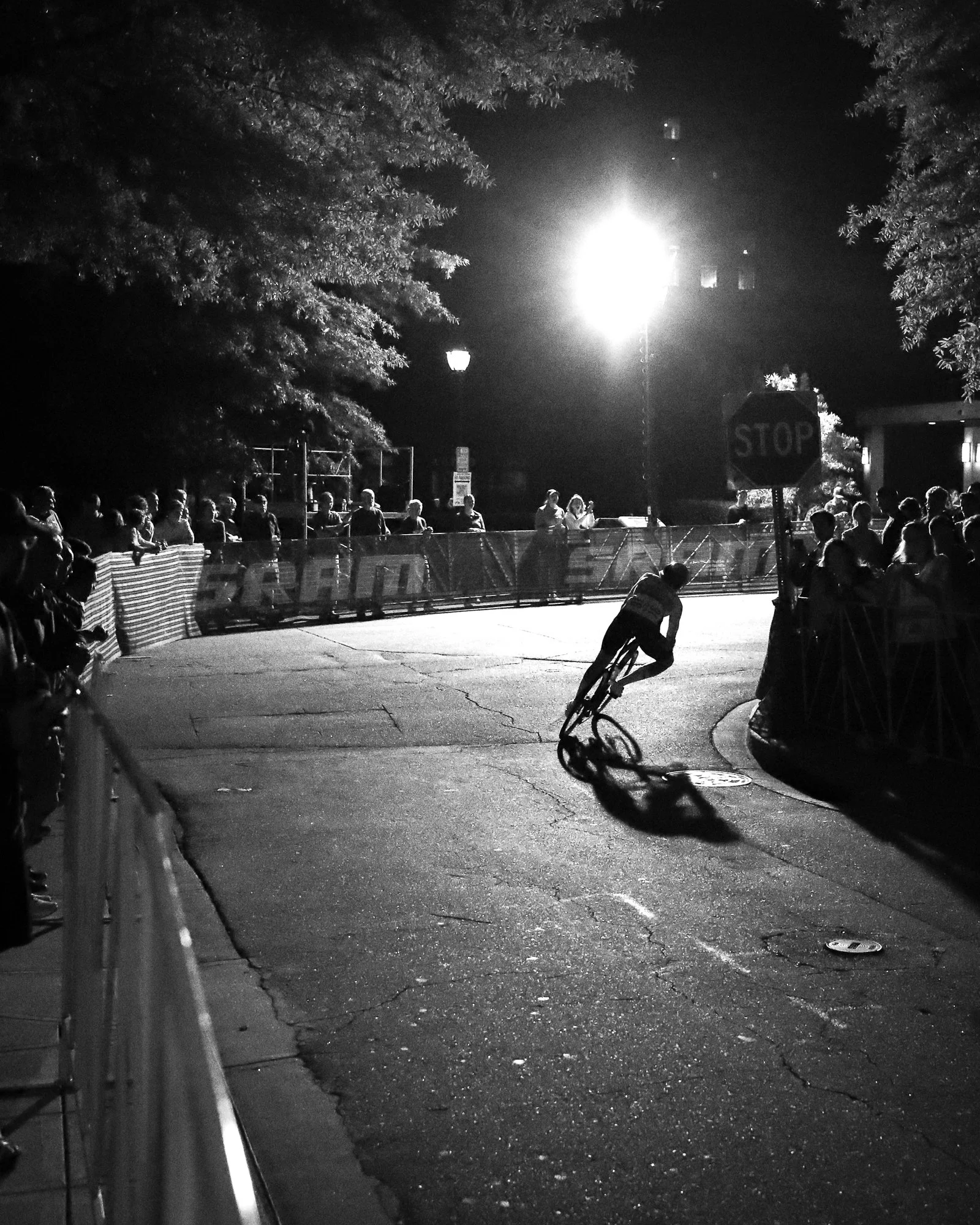 Nighttime scene of a group of people standing behind a barricade, with a cyclist in front of the barricade riding a bike on the street. Bright streetlight and trees visible in the background, along with a stop sign on the right side of the image.