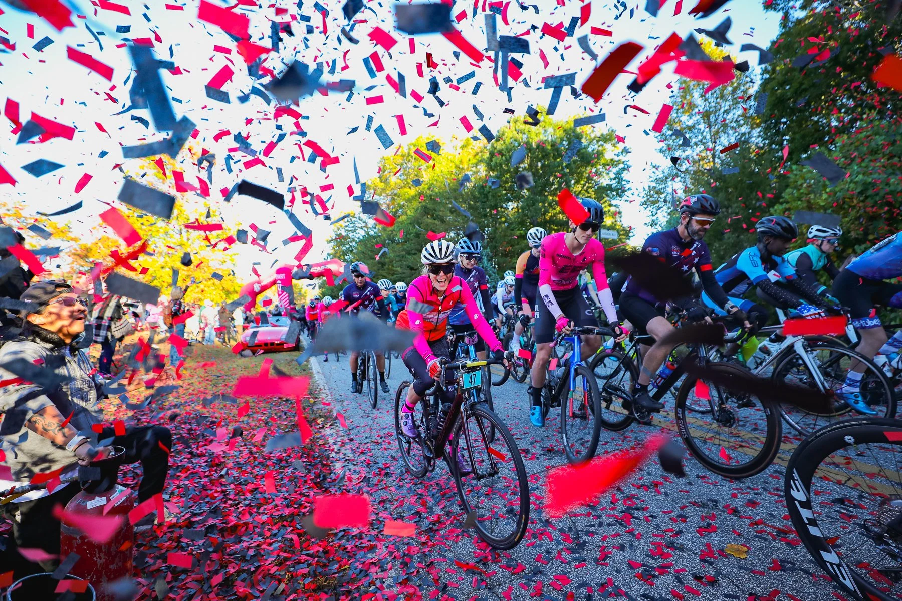 Cyclists participating in a race with red, black, and pink confetti falling around them and cheering spectators on the sidelines.
