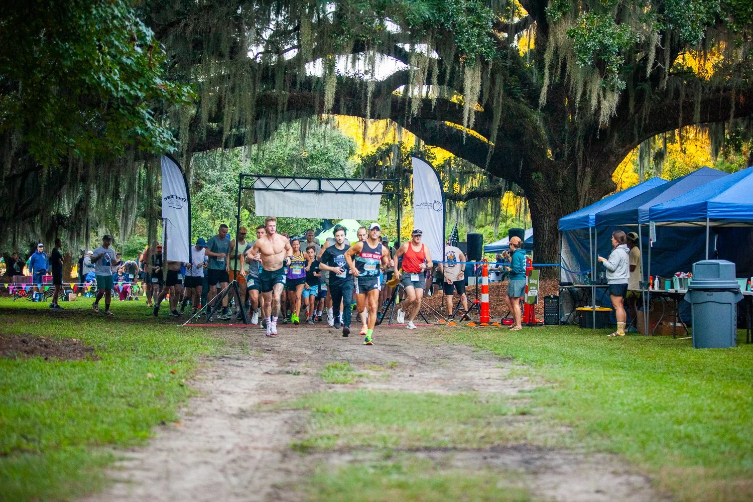 A group of runners start a race under a large tree with hanging moss, in a park setting, with aides and spectators nearby.