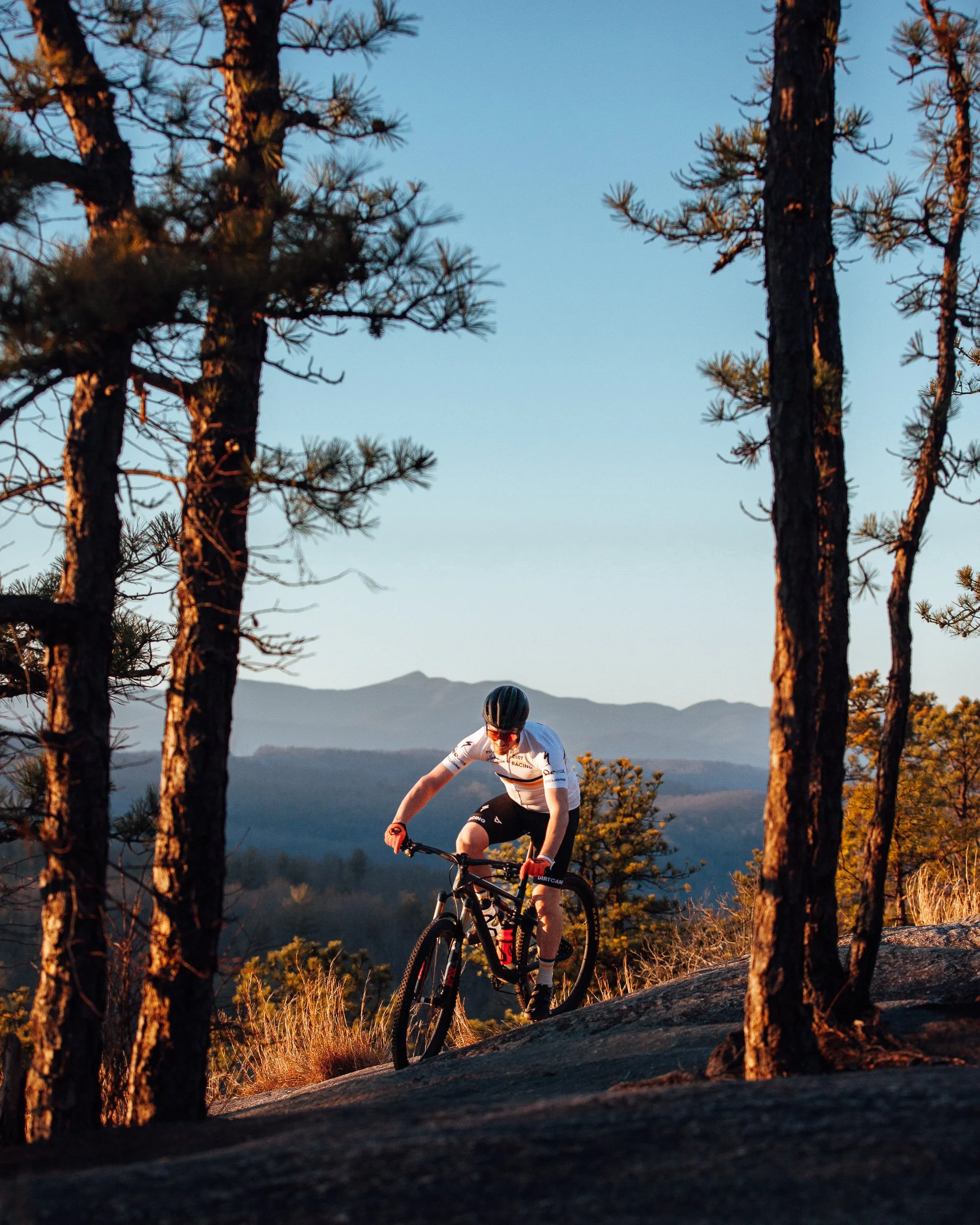 A mountain biker riding on a rocky trail surrounded by tall trees at sunset with mountains in the background.