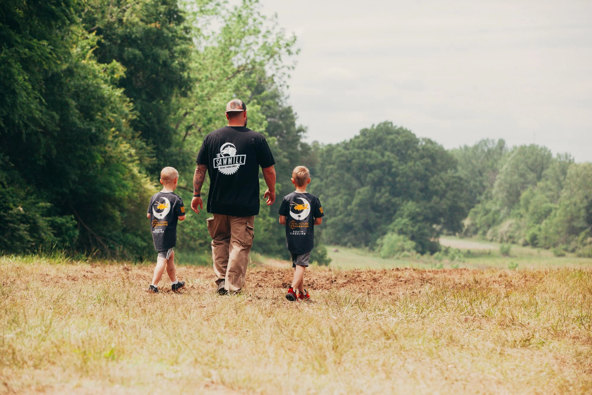 A man walking with two young boys outdoors in a grassy area with trees in the background, all wearing black shirts with a logo and text related to a seaplane foundation.