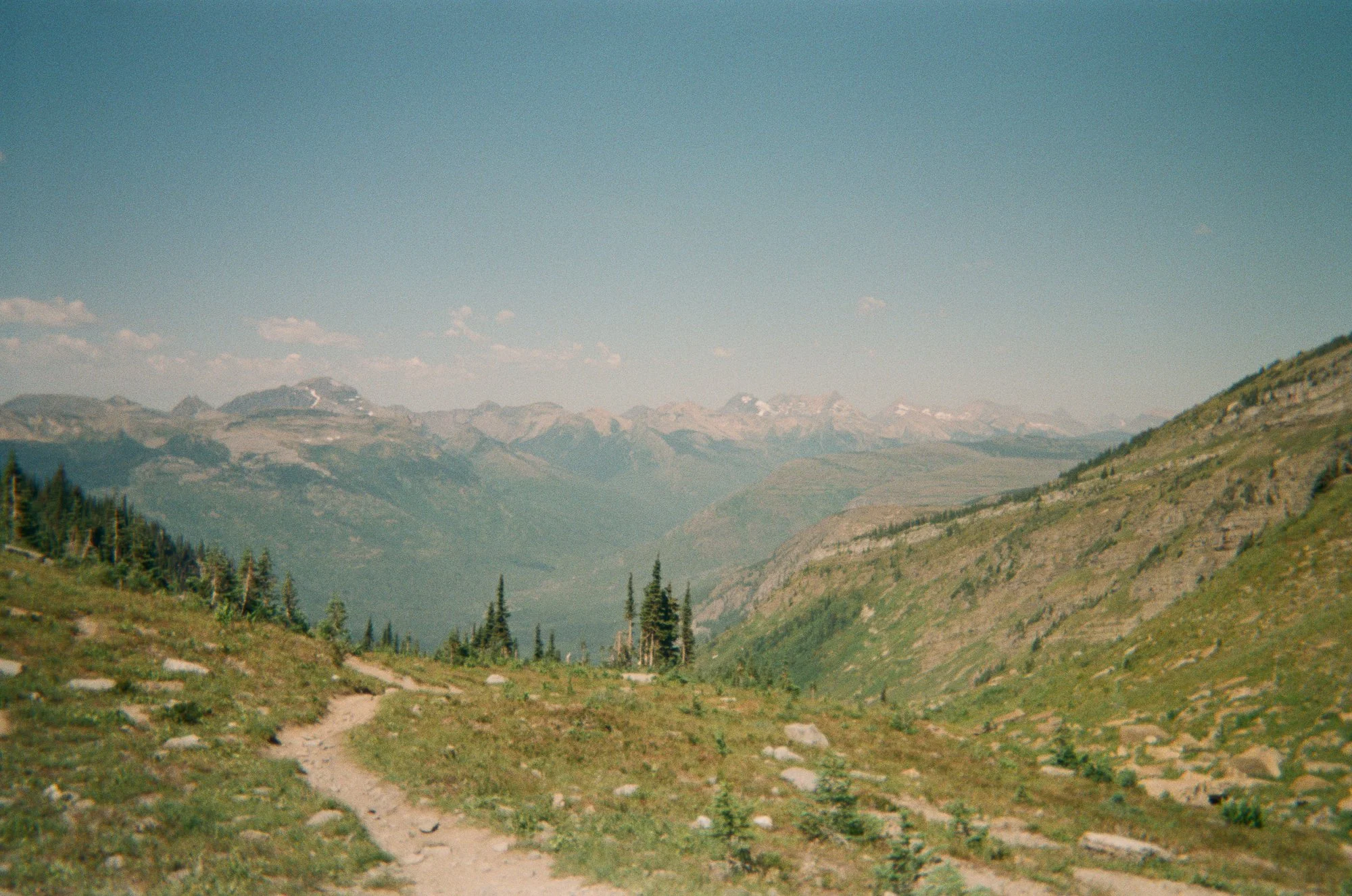 A mountain landscape with snow-capped peaks, a grassy valley, and a dirt trail in the foreground.