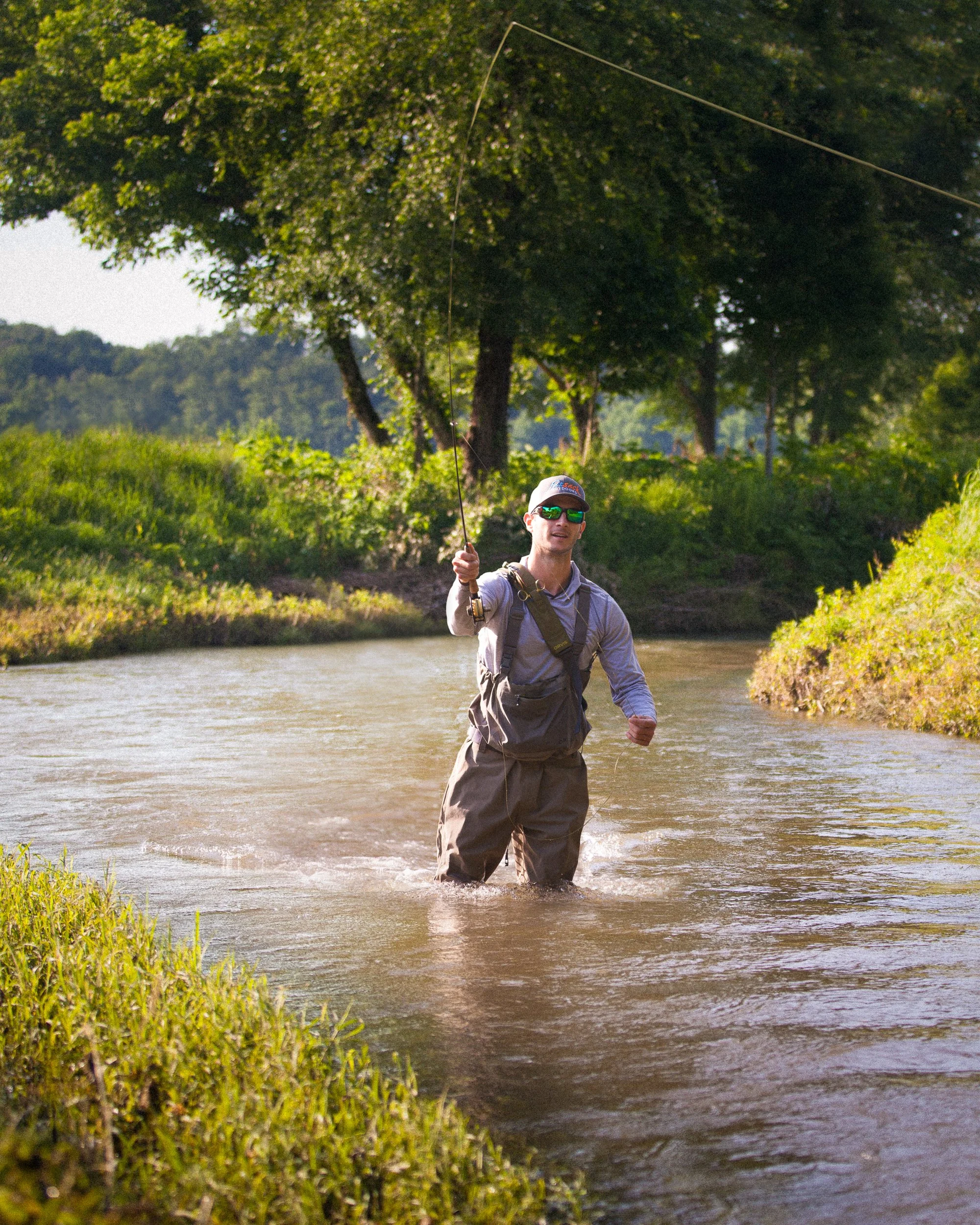 A man fishing in a river surrounded by green grass and trees, wearing sunglasses, a hat, and outdoor clothing.