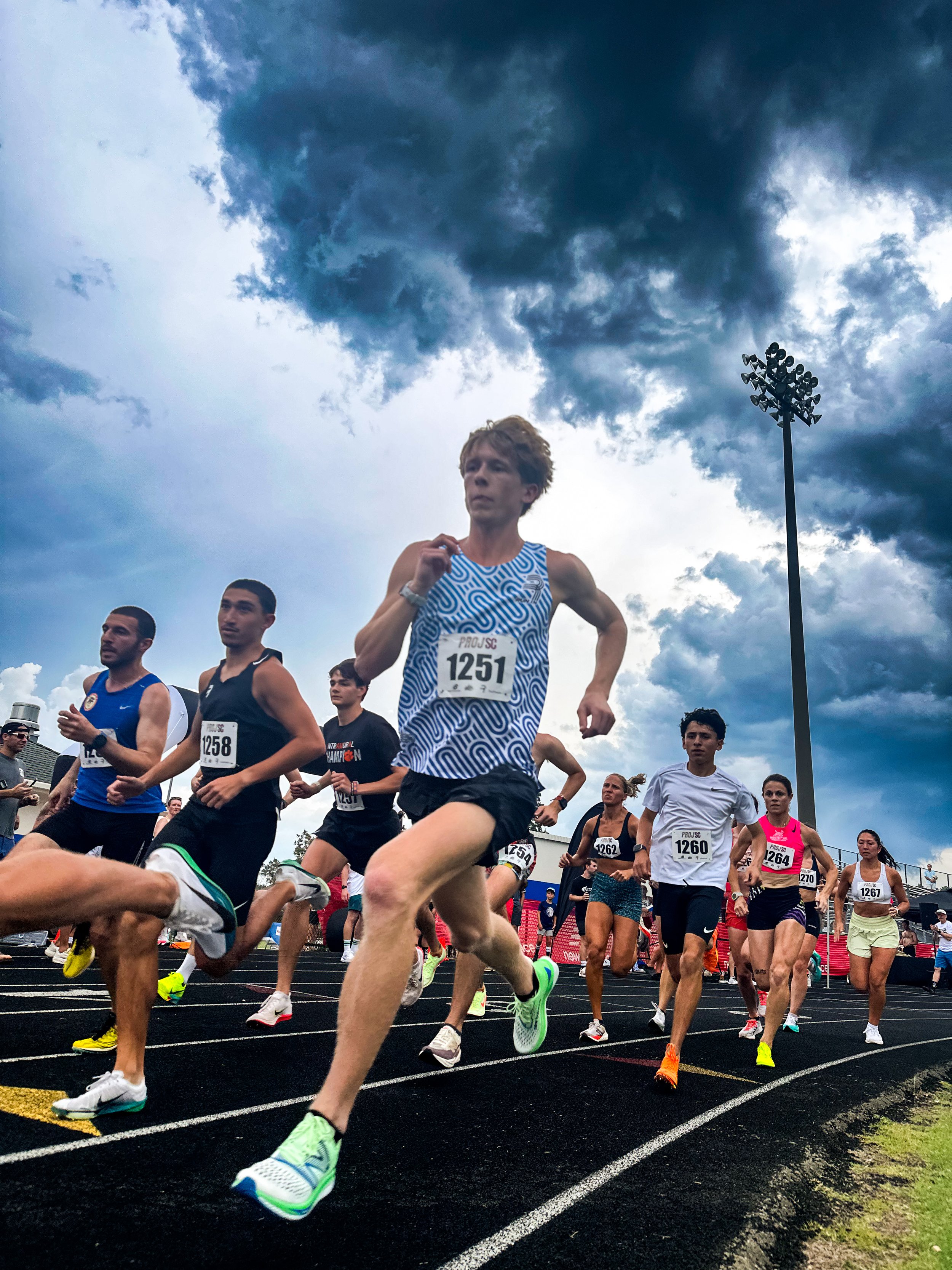 Group of runners participating in a race on a dark track under a cloudy sky, wearing athletic gear with race bib numbers.