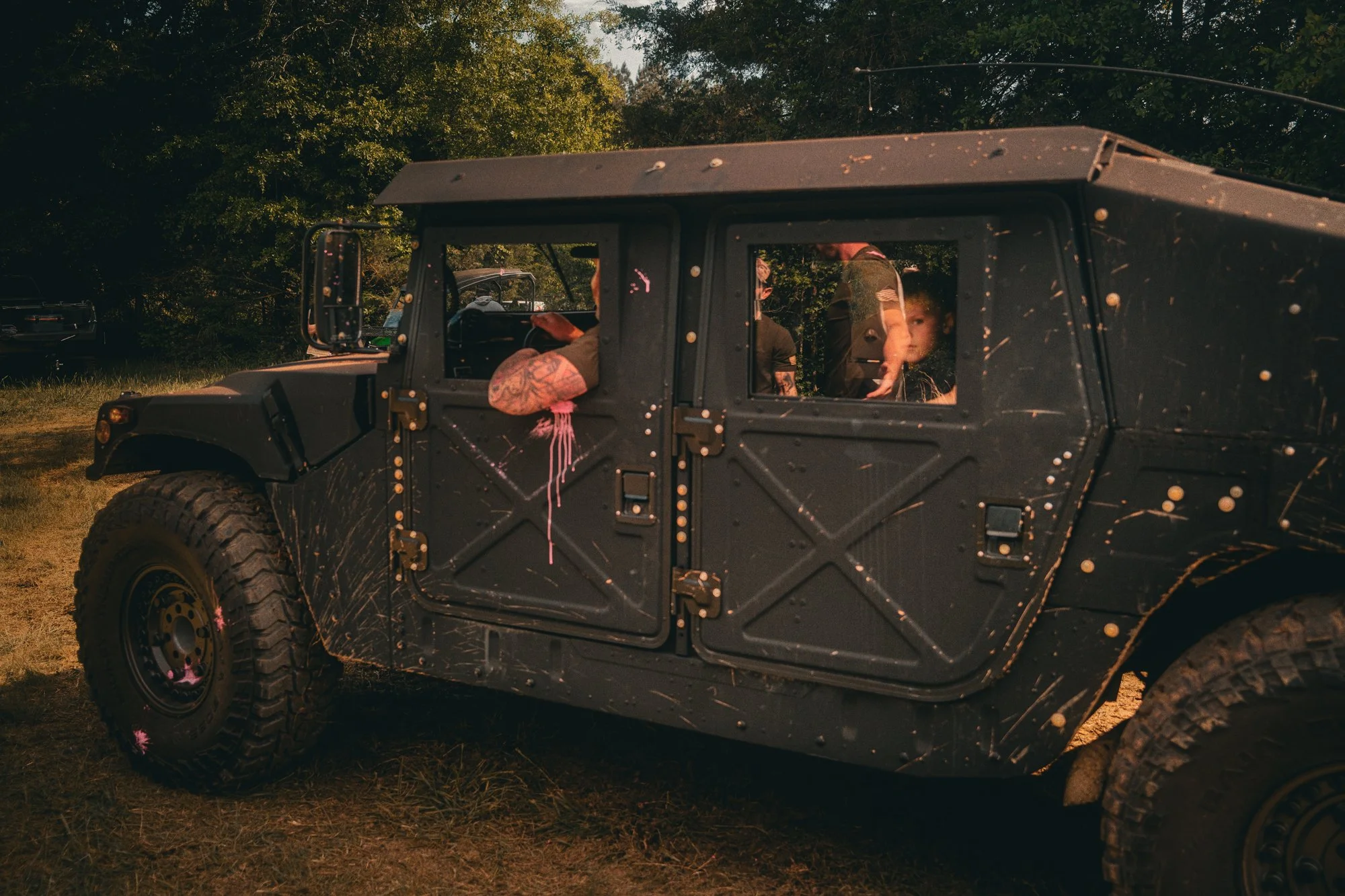A black military-style vehicle with four doors and large rugged tires, parked outdoors on grass with trees in the background. Inside the vehicle, children and adults are visible through the windows, some looking out and others engaged in activities.