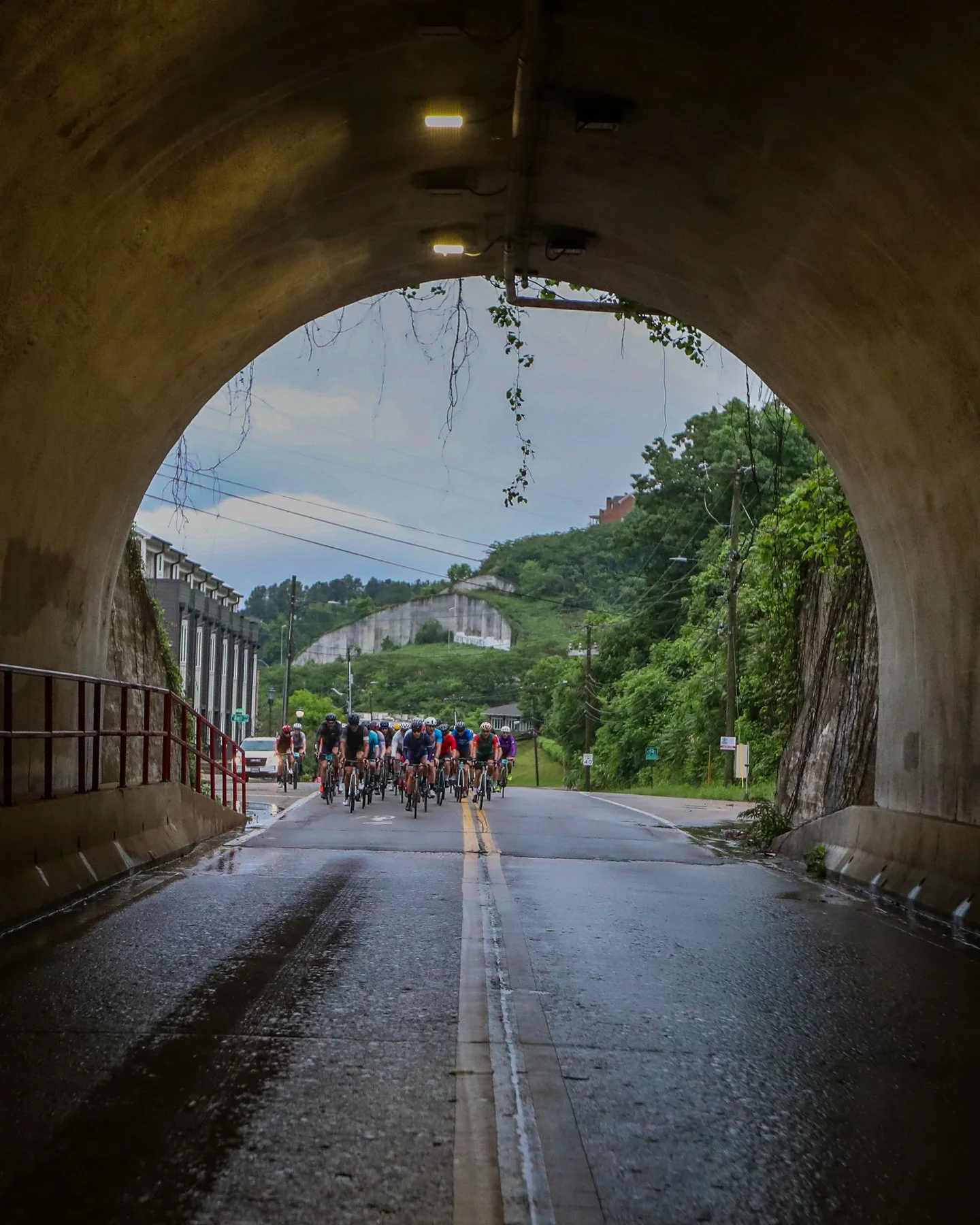 View from inside a tunnel looking out at a group of cyclists riding on the road. The tunnel has a curved ceiling with lights, and there are trees and a hillside visible in the background.