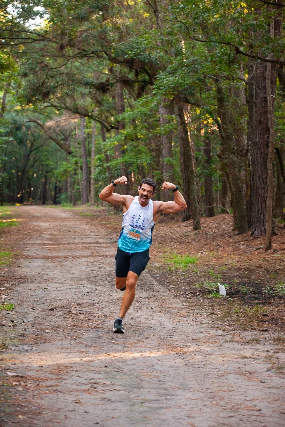 A man running on a dirt trail in a forest, holding his arms up in victory, smiling, wearing a sleeveless shirt and shorts, with a race number tag on his chest.