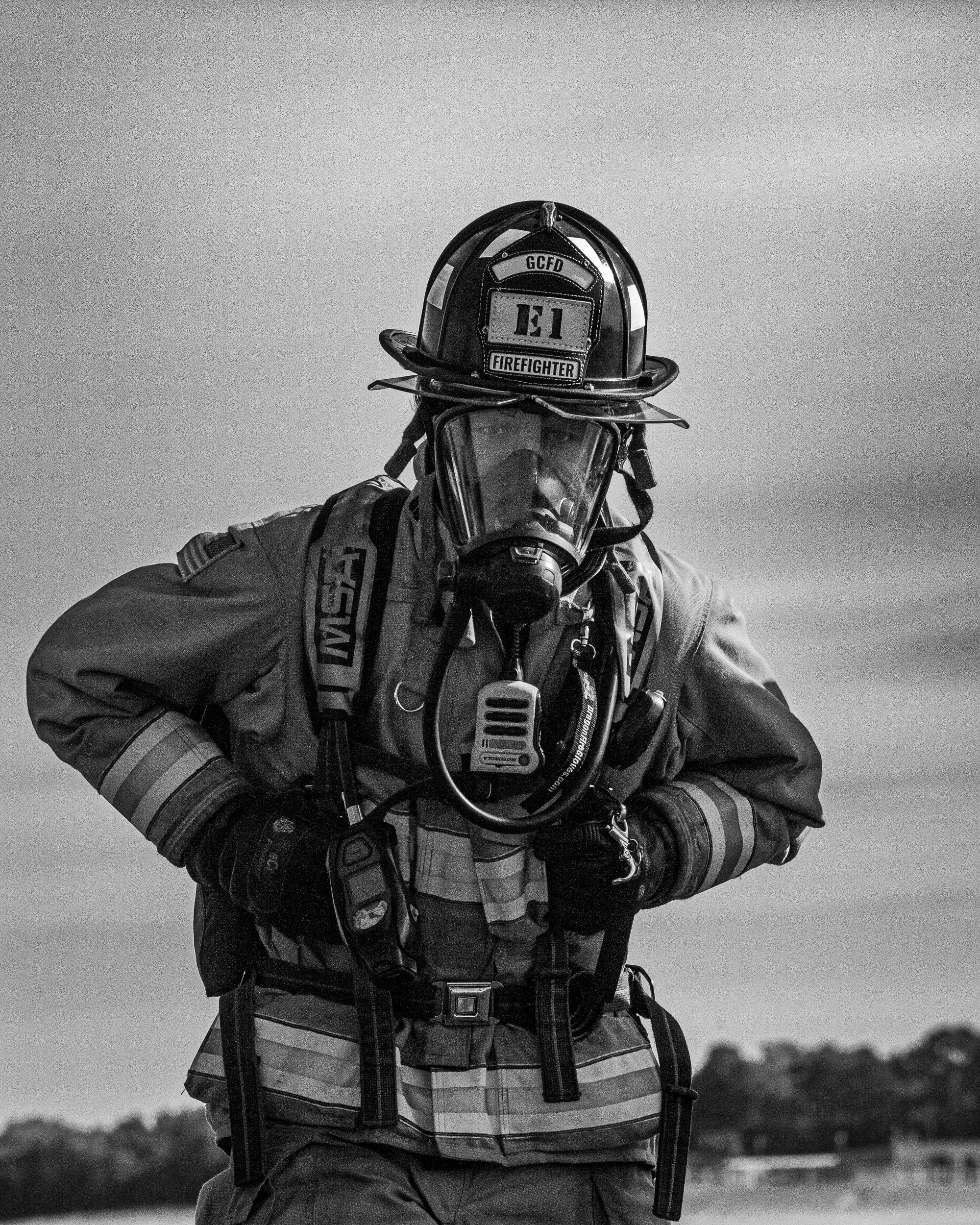 Black-and-white photo of a firefighter pointing towards the camera, wearing a helmet with a badge that has 'FIRE FIGHTER' and a patch with 'GCFD' and logo, a protective mask, and gear, standing outdoors with trees in the background.
