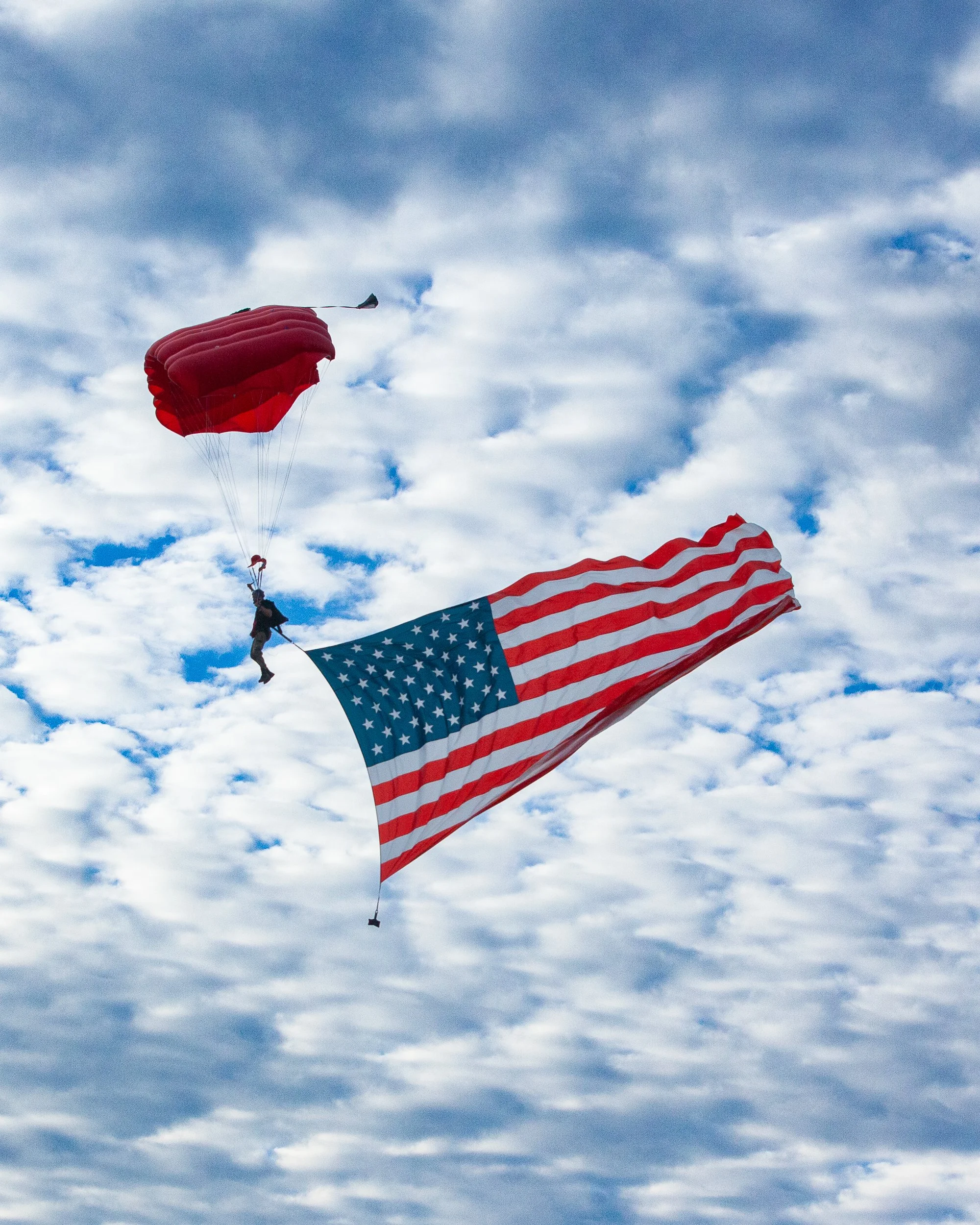 A person flying with a parachute in the sky, accompanied by a large American flag waving in the wind against a backdrop of clouds.