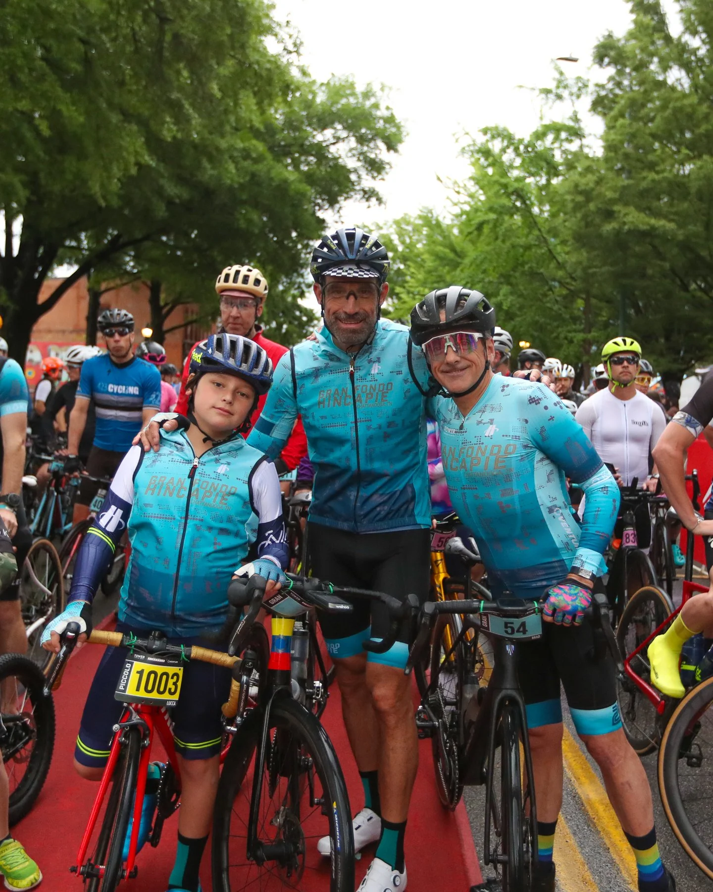Three cyclists in matching blue jerseys, preparing for a race, with other participants and trees in the background.