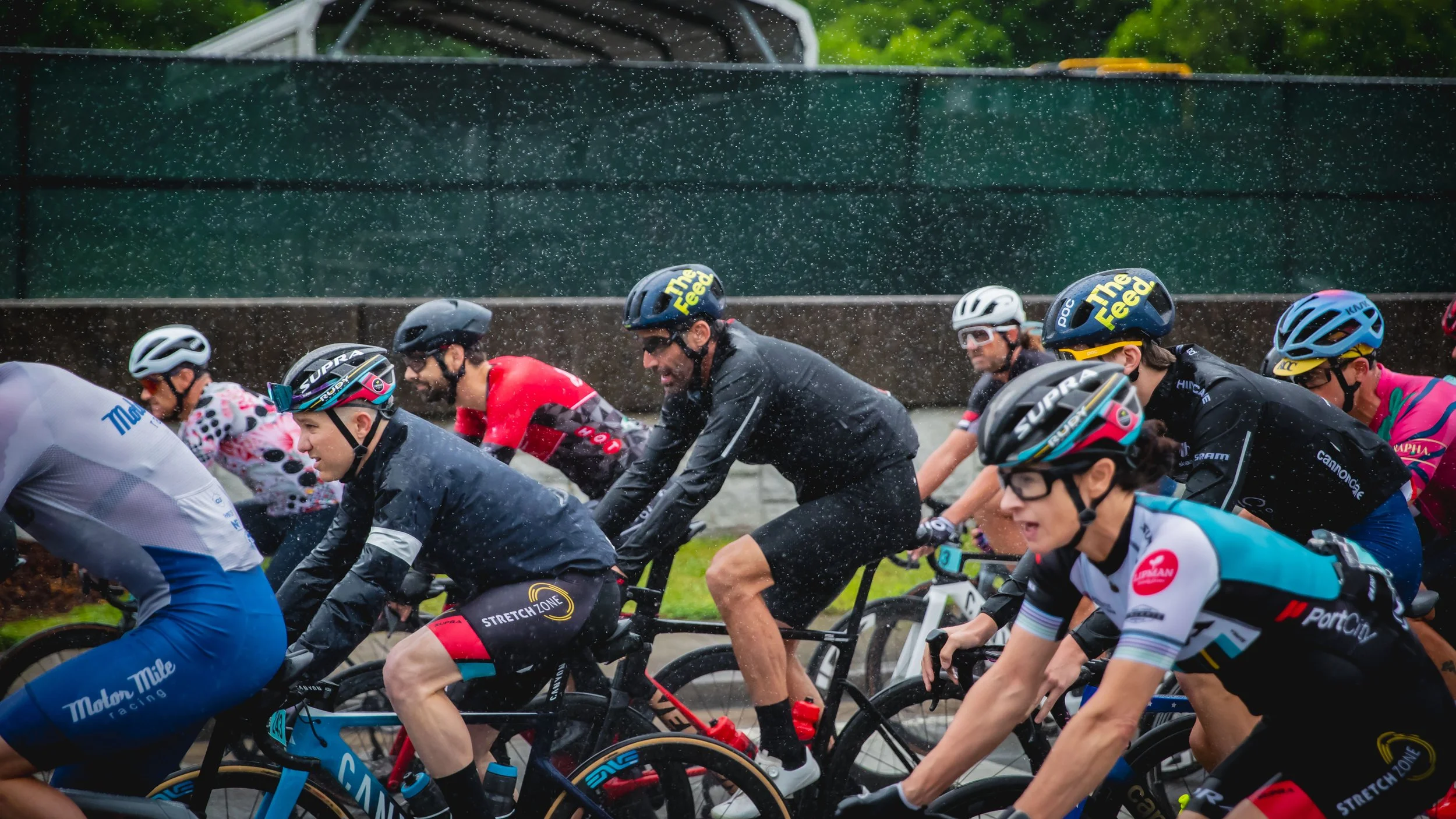 Cyclists participating in a race, riding their bikes under rainy weather with raindrops visible, wearing helmets and colorful racing gear.