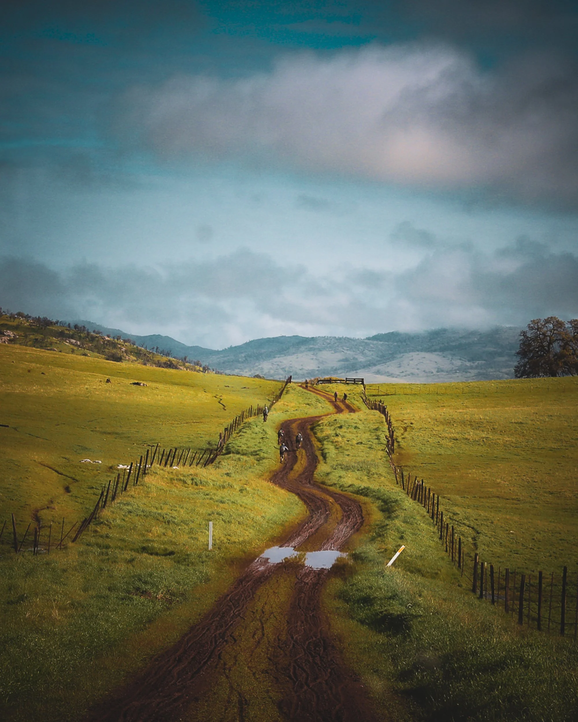 Dirt road winding through green fields with fences, leading to hills and mountains under a cloudy sky.