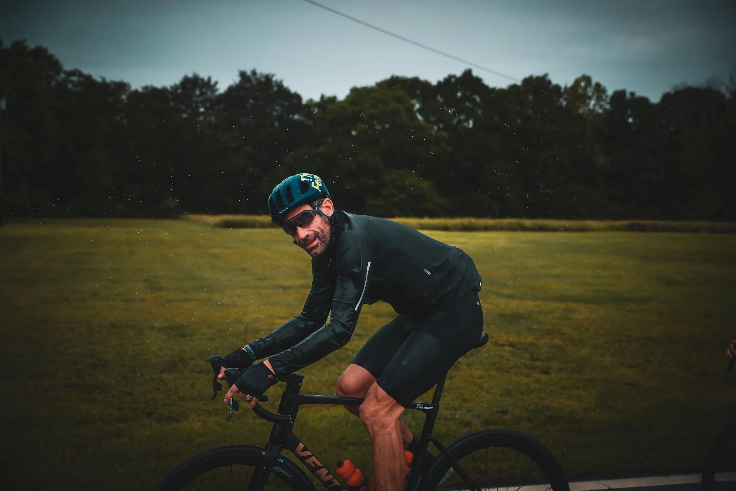 A man wearing a helmet and sunglasses riding a bicycle outdoors in a grassy field with trees and a cloudy sky in the background.