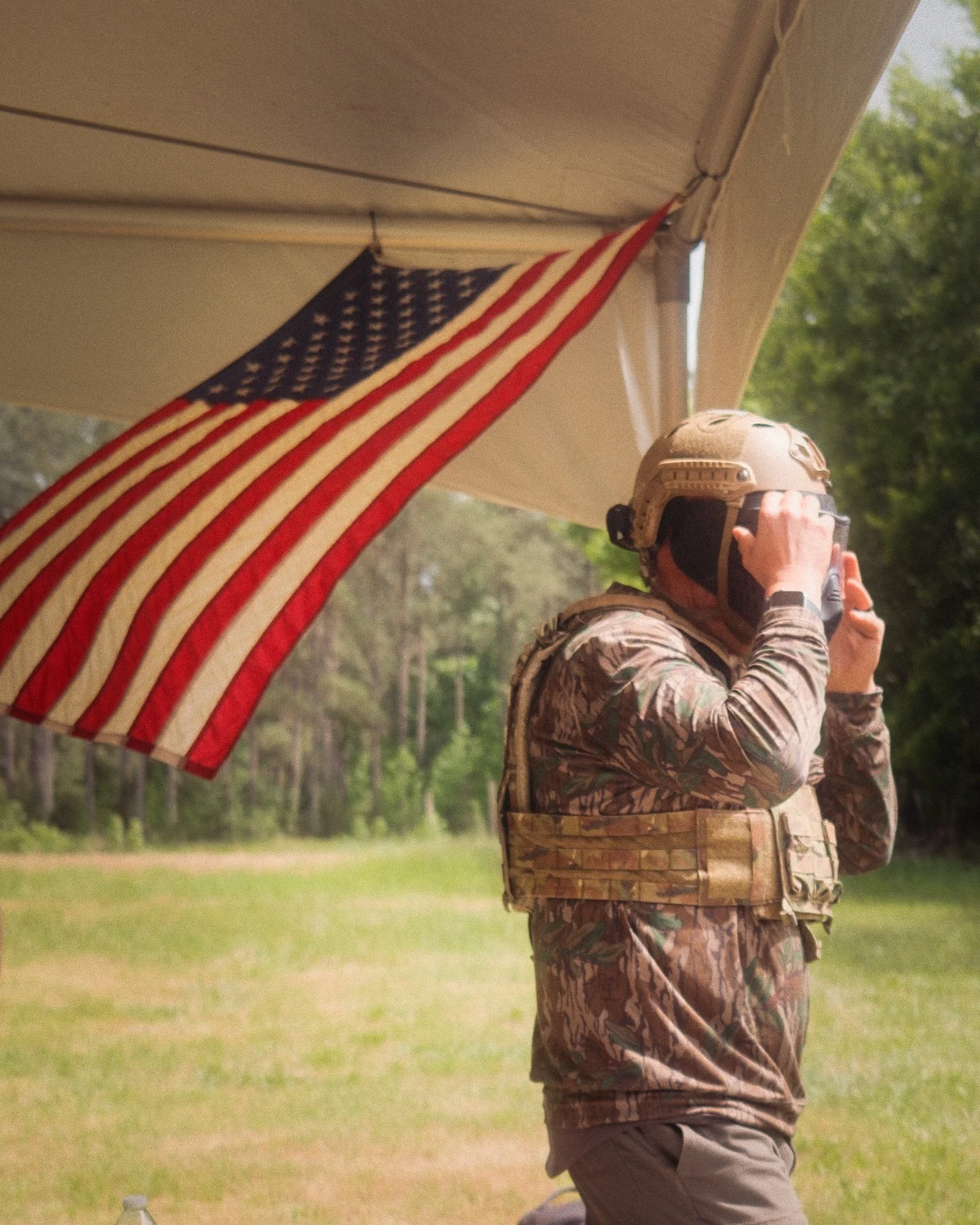 A soldier in camouflage uniform with a helmet adjusts his goggles under an American flag hanging from a wooden structure outdoors.
