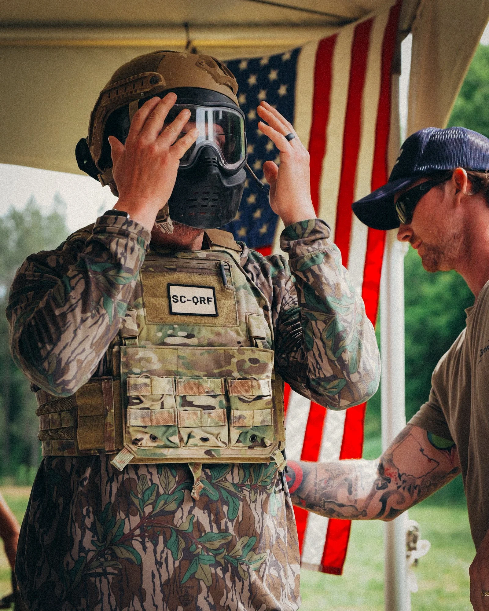 A person in military camouflage uniform places a helmet and goggles on their head at an outdoor event. Another person with tattoos and a baseball cap is partially visible. An American flag hangs in the background.