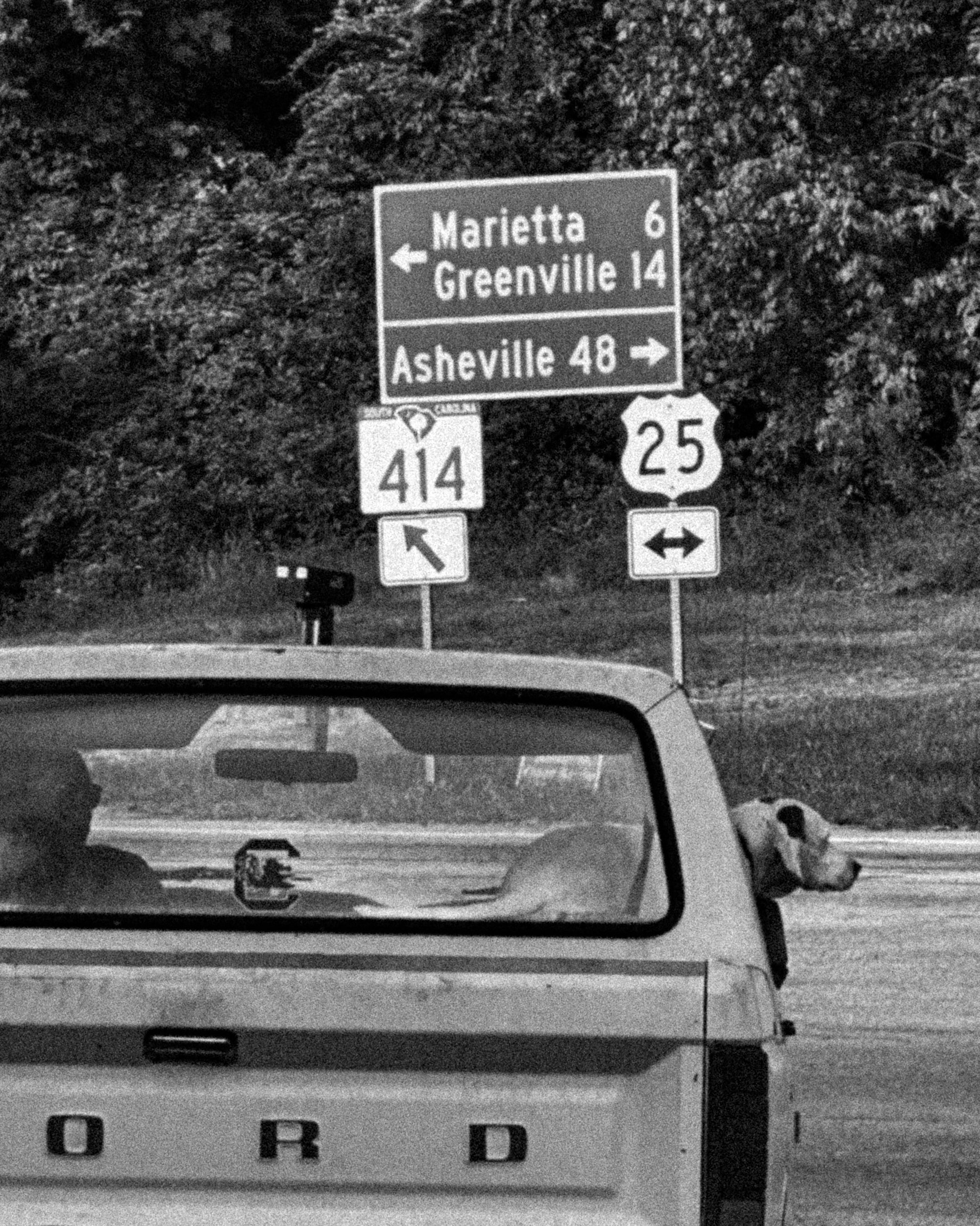 Black and white photo of road signs at night, including directions to Marietta, Greenville, and Asheville, with trees in the background and a car visible below.