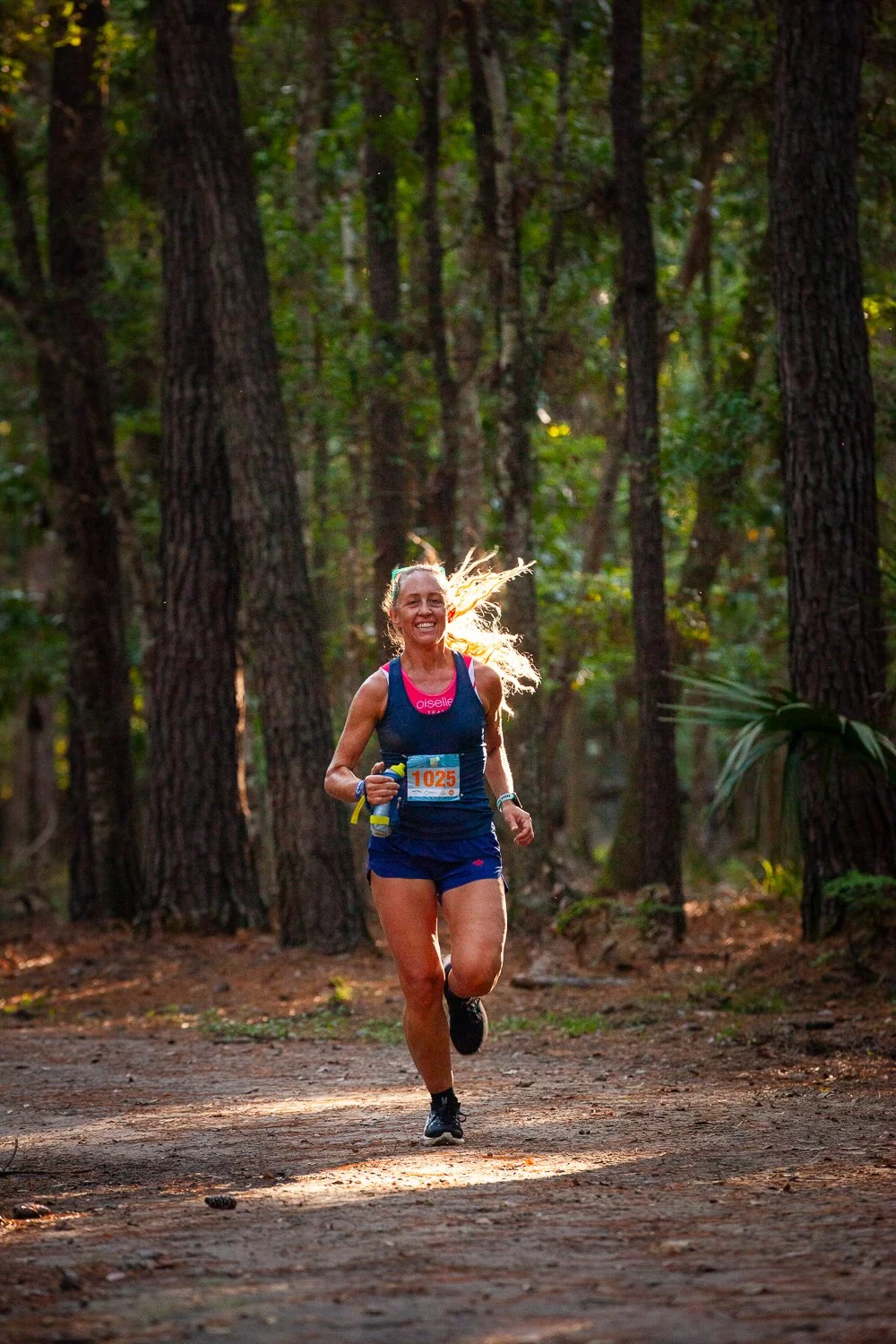 A woman running on a dirt trail through a forest, smiling with sunlight shining behind her.