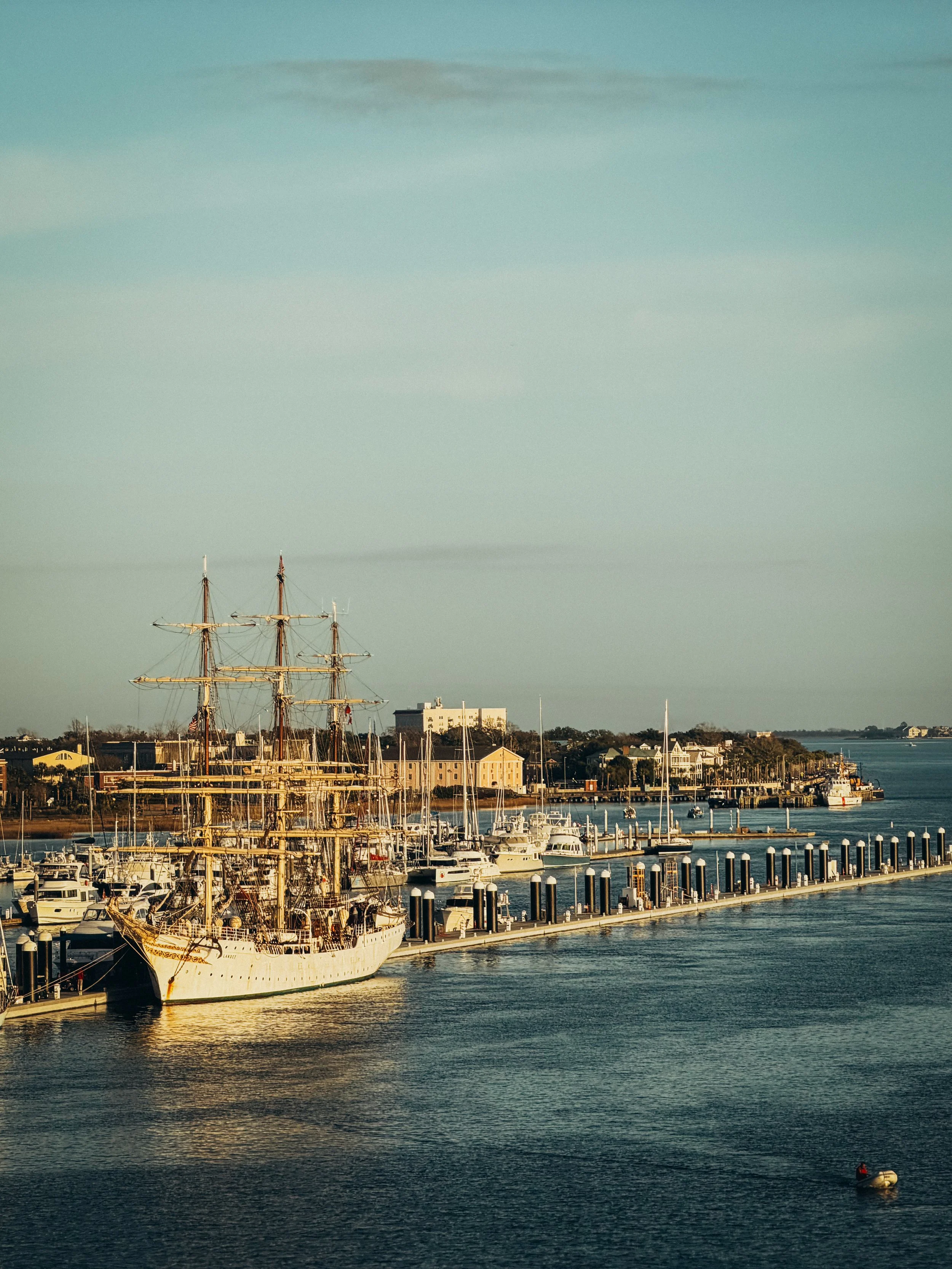View of a harbor with a large, historic sailing ship anchored at the dock, surrounded by smaller boats and yachts, with buildings and trees along the waterfront under a partly cloudy sky.
