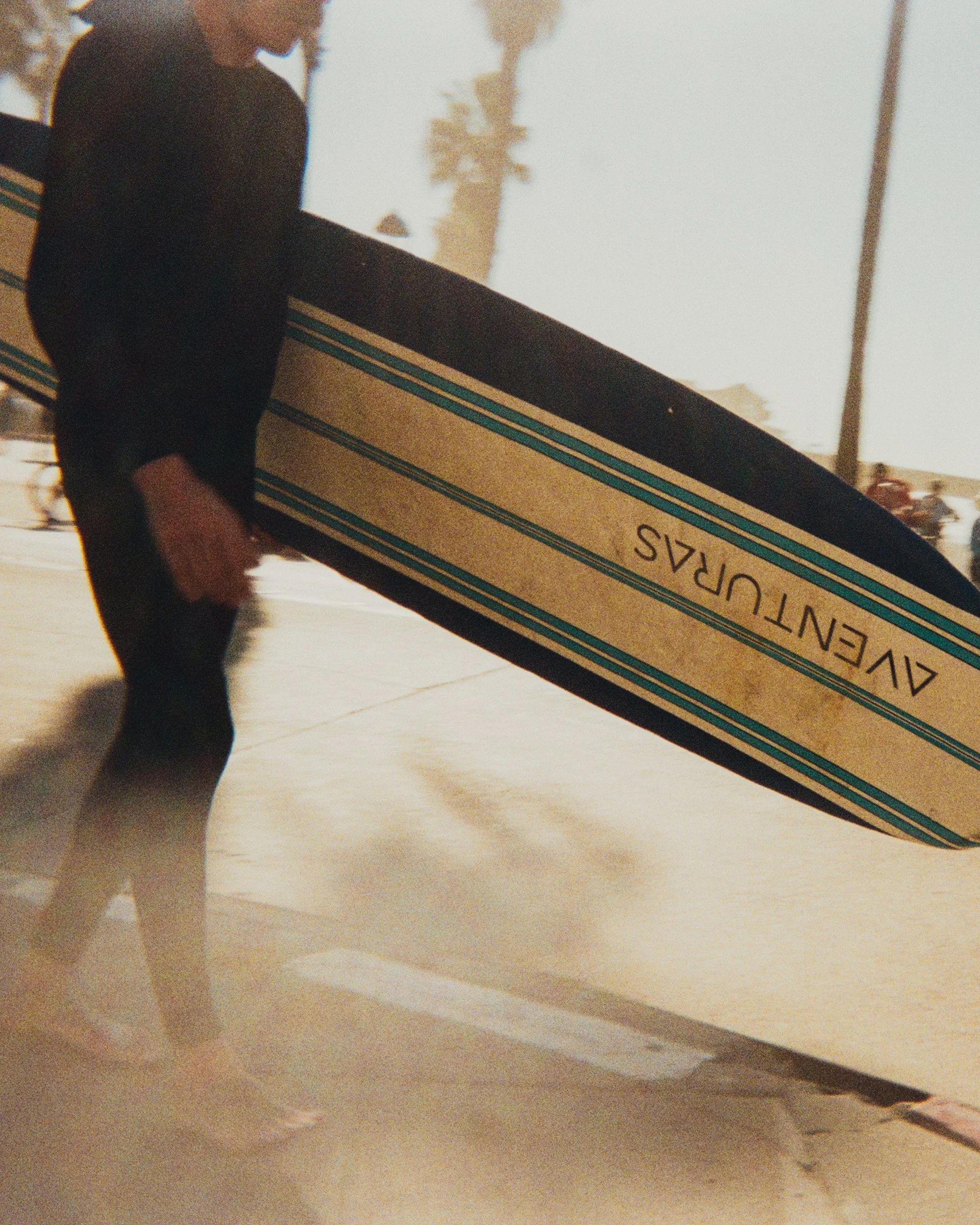 Person holding a long rectangular box, standing on a skateboard, outdoors with palm trees in the background.