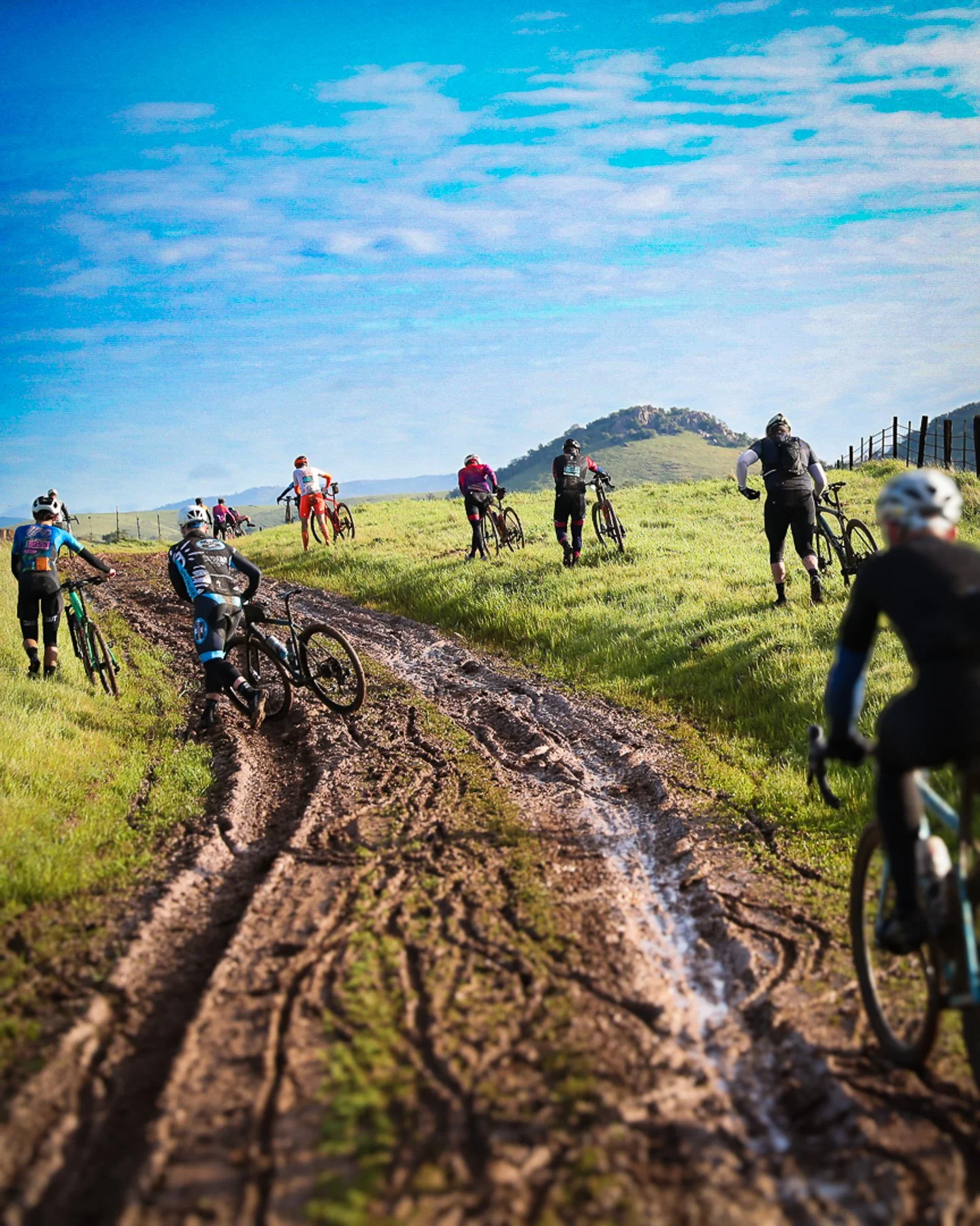 Group of mountain bikers riding on a muddy trail through green fields with hills in the background under a blue sky.