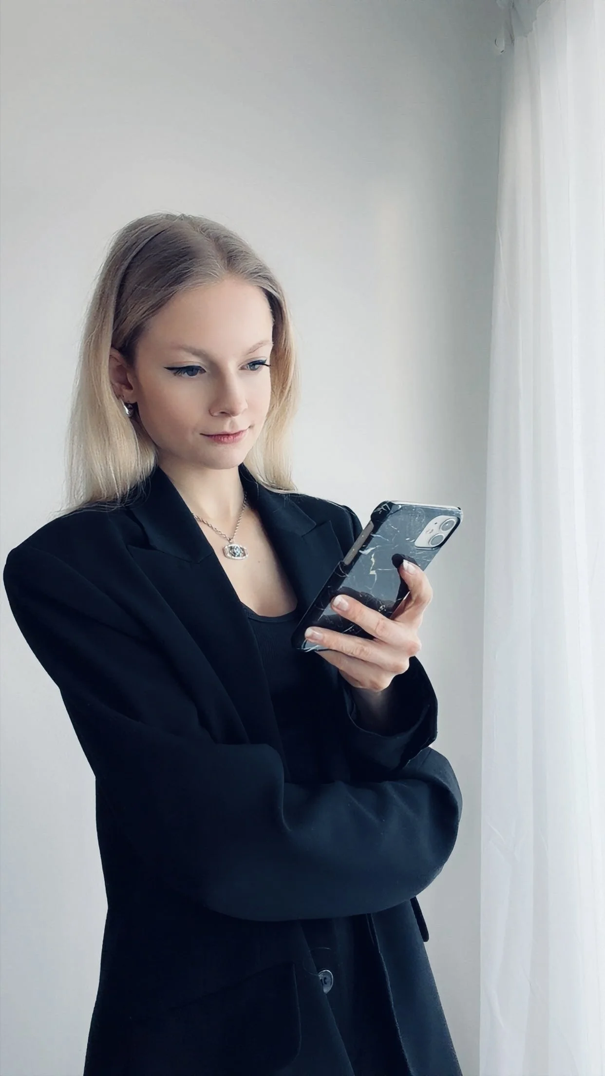 Blonde Frau mit schwarzem Anzug blickt lächelnd auf ihr Handy in der Hand.