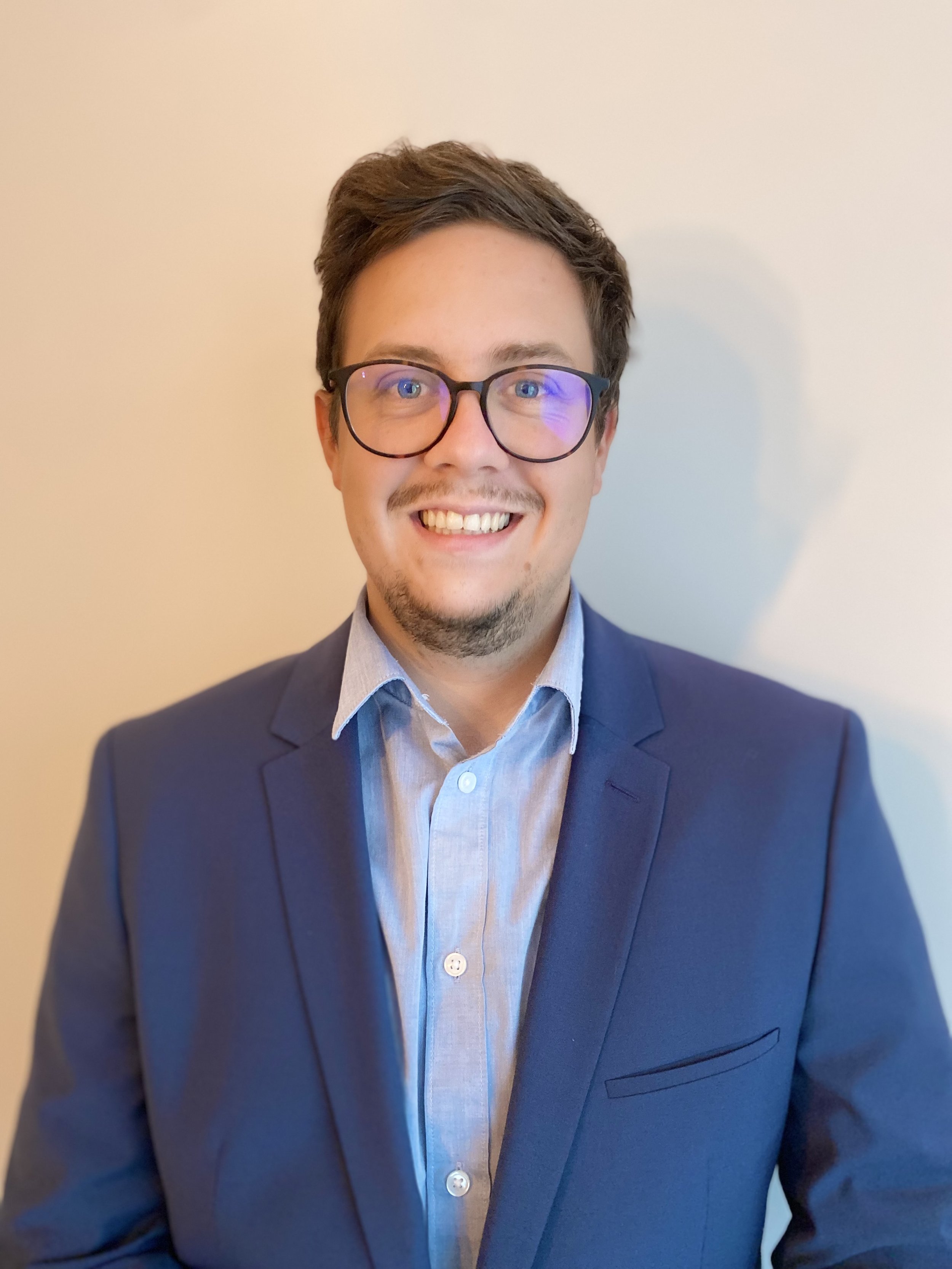 Mann mit Brille, braunen Haaren und blauem Anzug lächelt gerade in die Kamera.