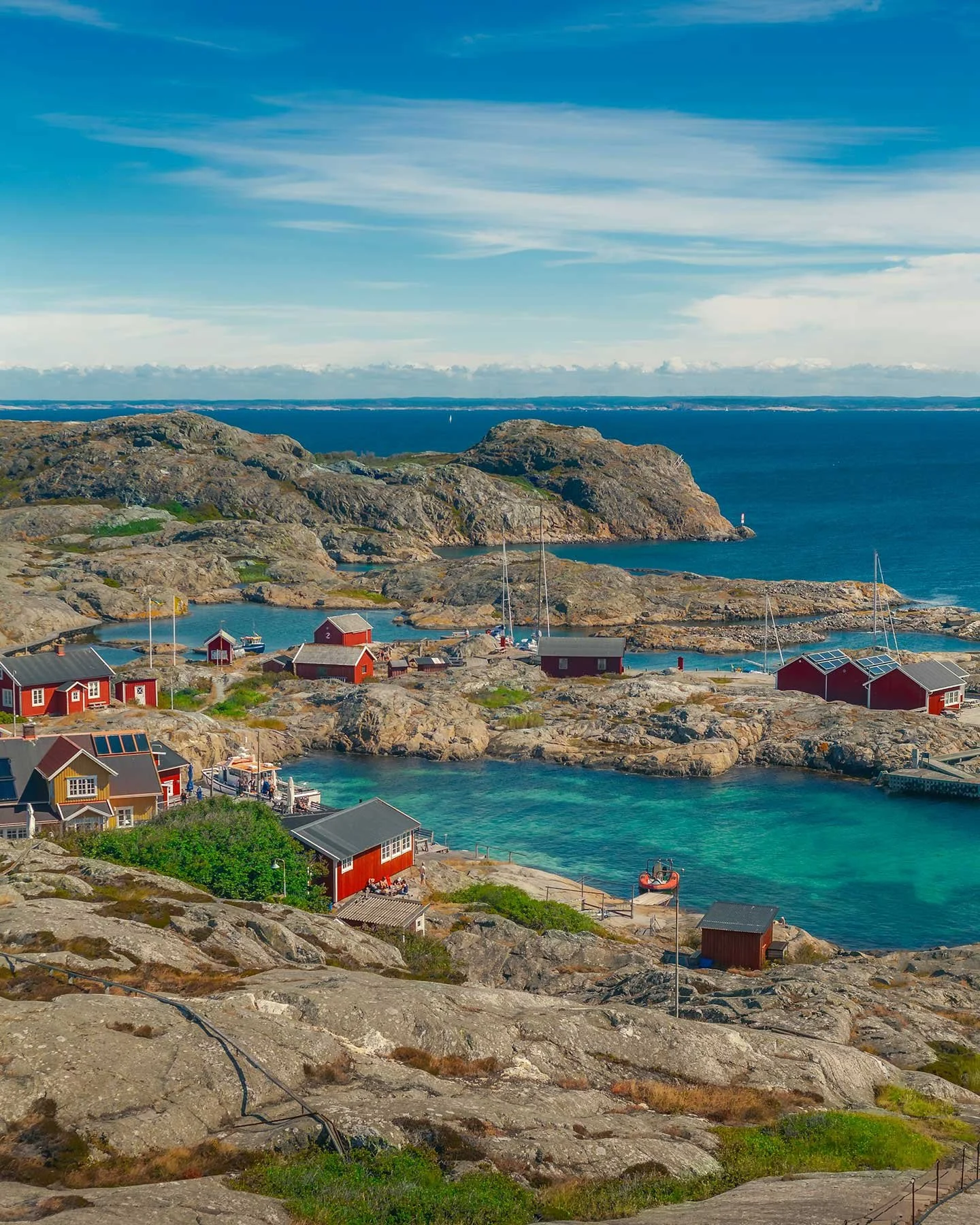 Fjällböckar med röda hus vid havet, båtar och klippor under en blå himmel.