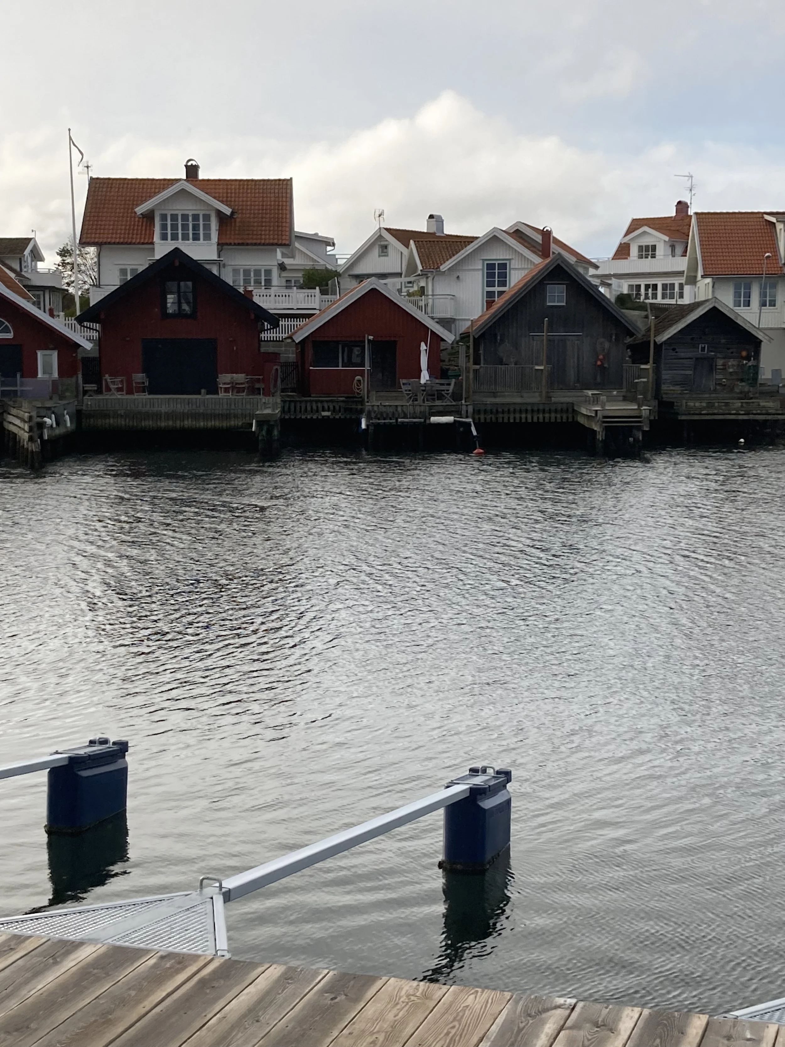 Flera hus längs med vattnet, några är vita med röda tak, andra är mörka med röda tak. Även en liten brygga och vatten är synligt.
