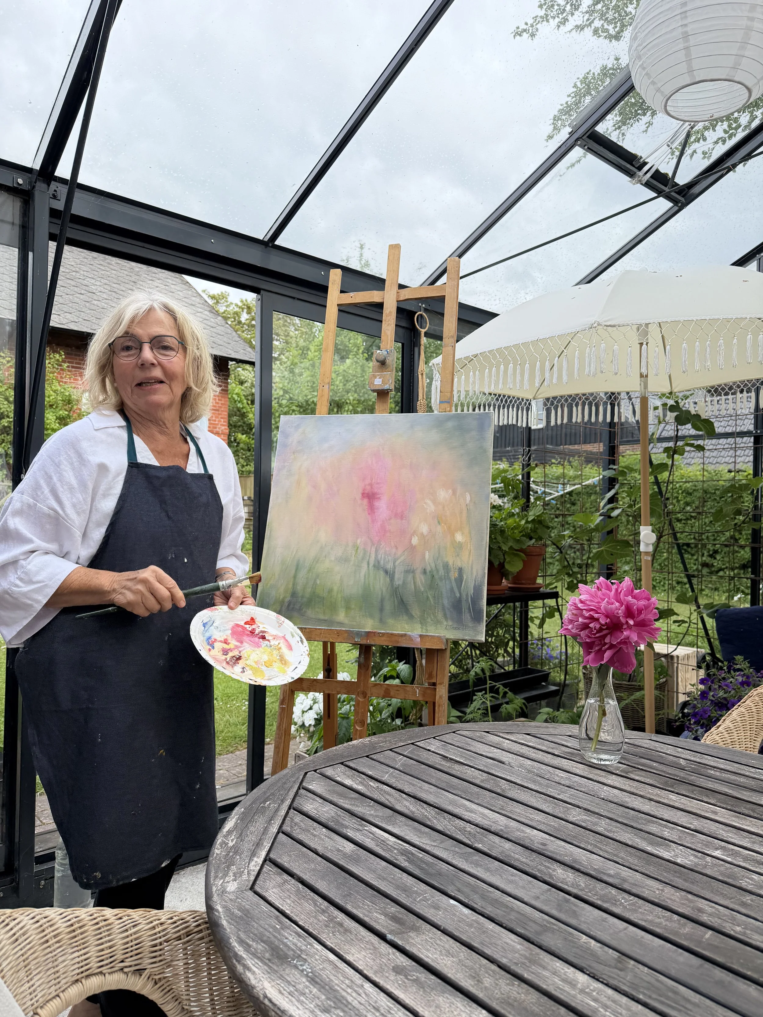 En kvinna som målar en abstrakt solnedgång på staffli i ett växthus med hängande lampor och blommor på bordet, en växtkruka och en parasoll syns.