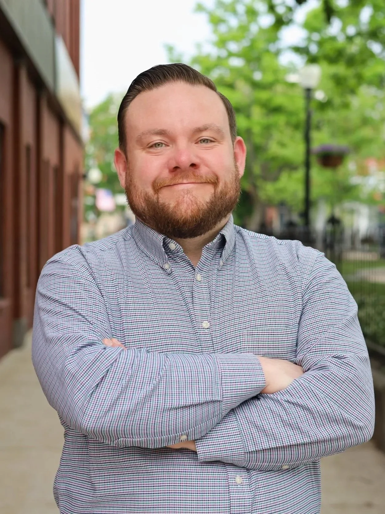 Nick Paul smiling with arms crossed, standing outdoors on a sidewalk with trees, a lamppost, and a brick building in the background.