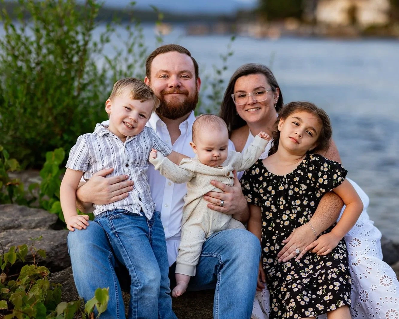 A family of five sitting outdoors near Lake Winnipesaukee, smiling at the camera. The group includes two adults and three children, with greenery in the background.