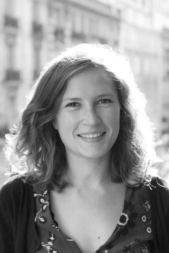 Photo en noir et blanc d'une femme souriante avec des cheveux bouclés, portant un haut à motifs floraux, avec un bâtiment en arrière-plan