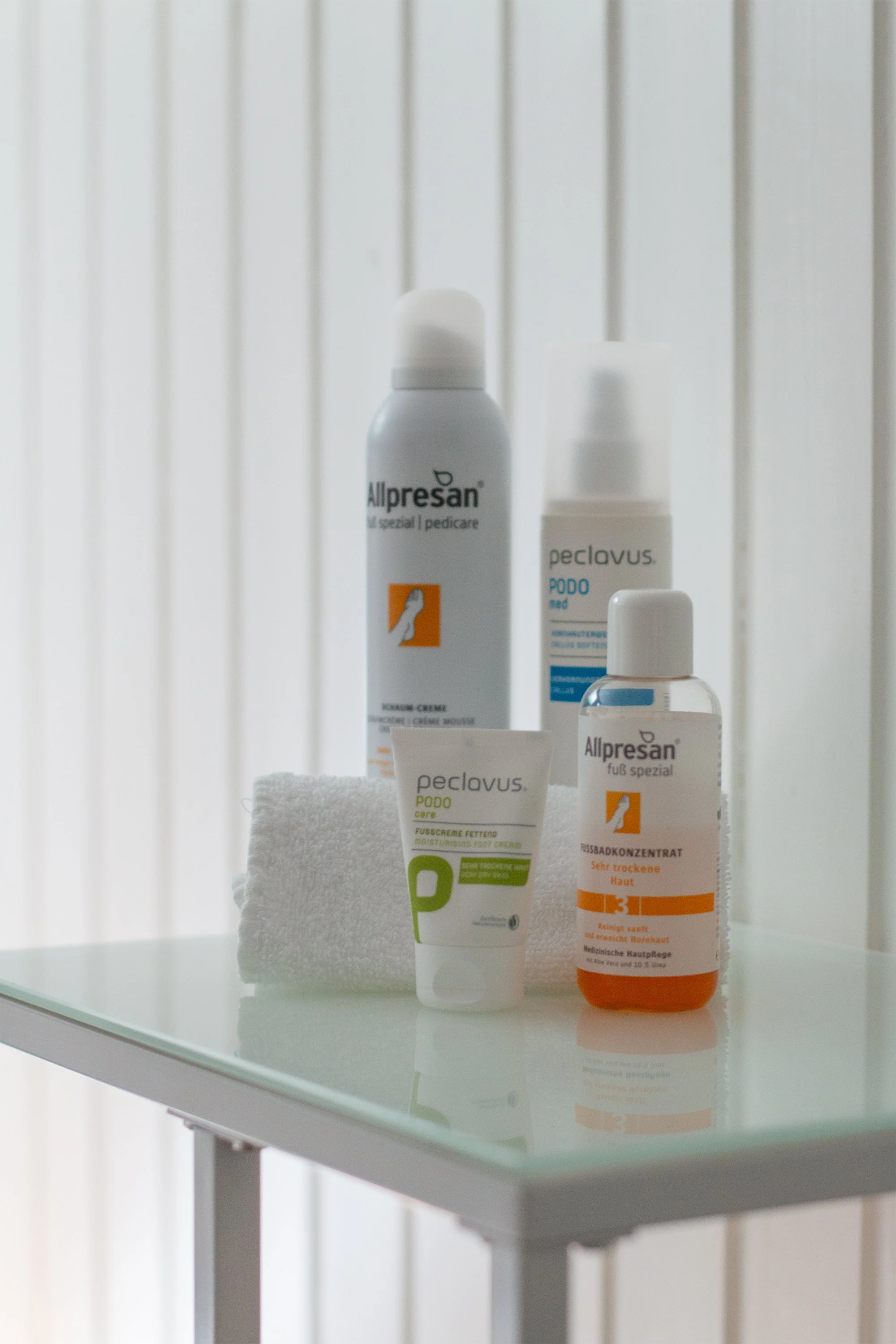 Pflegeprodukte auf einem weißen Tisch, inklusive Allpresan Creme, Peclavus Fußcreme und Fußbadkonzentrat, mit weißen Handtuch und einer weißen Oberflächen im Hintergrund.