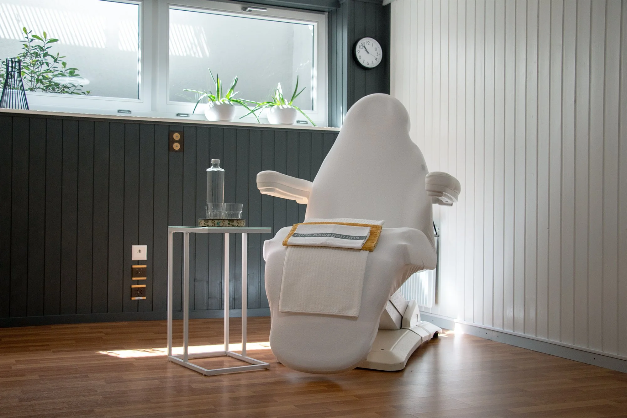 Ein modernes Behandlungszimmer mit einem speziellen, weichen Stuhl, einem kleinen Beistelltisch mit Wasserflasche und Glas, einer Fensterbank mit Pflanzen und einer Uhr.