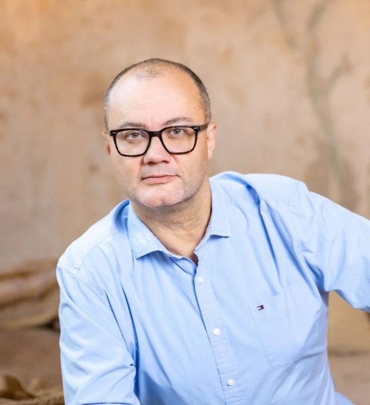 A bald man wearing black-rimmed glasses and a light blue button-down shirt with a small logo on the pocket, posing in front of a textured, neutral-colored background.