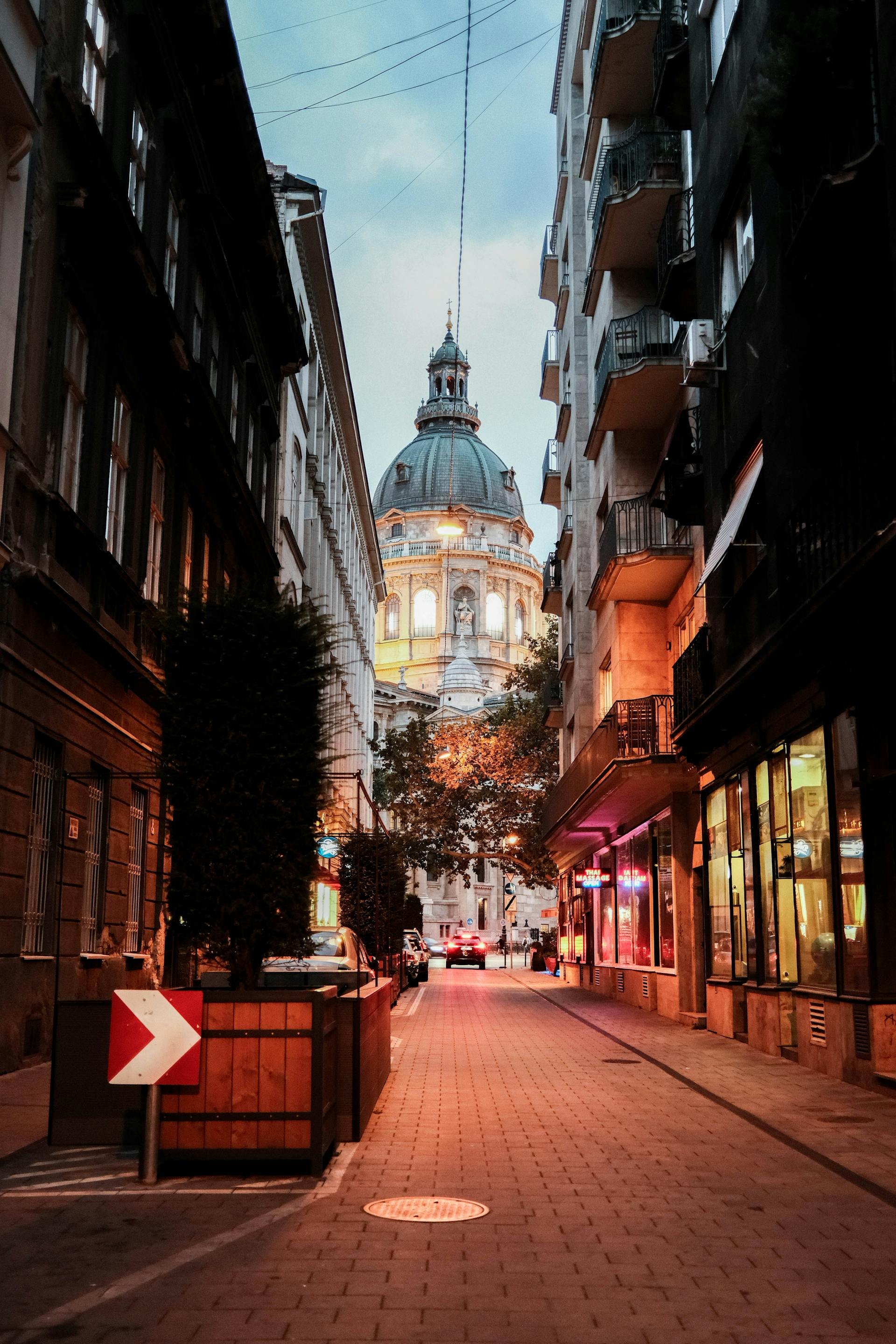 A narrow city street at dusk with a historic domed building in the background, illuminated by streetlights. The street is lined with multi-story buildings with balconies on the right and empty on the left. Cars are parked along the street, and a few are driving away. There are trees and illuminated signs on the right side of the street.