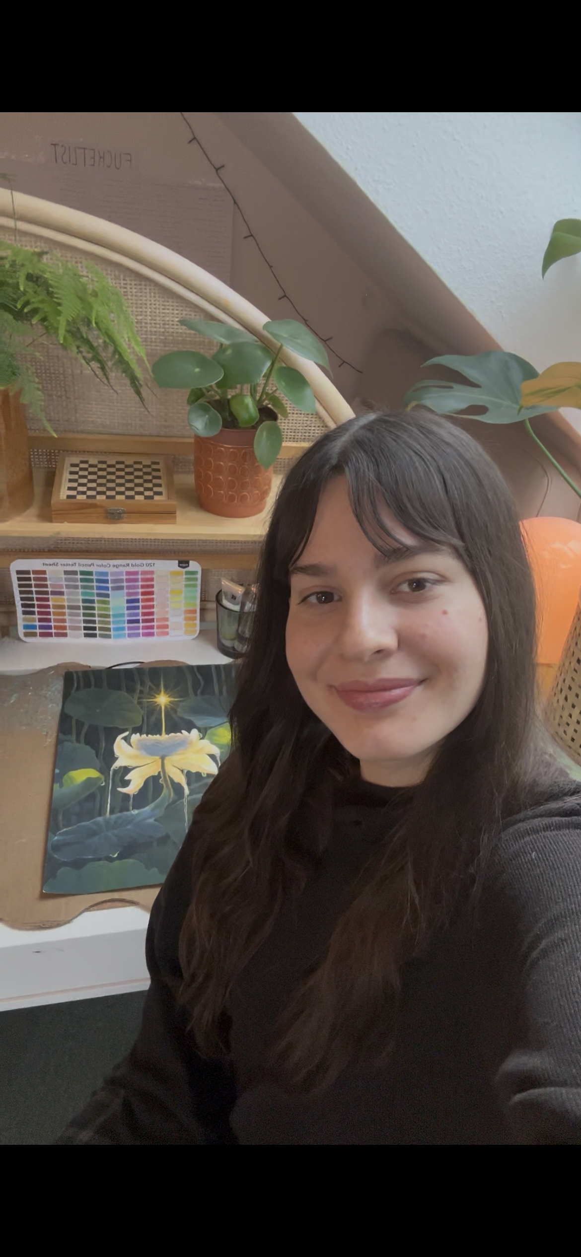 A young woman with long dark hair and wearing a black shirt, taking a selfie in a room with plants in the background, a colorful palette of paint swatches, and a painting of a yellow flower with a glowing center on the desk.