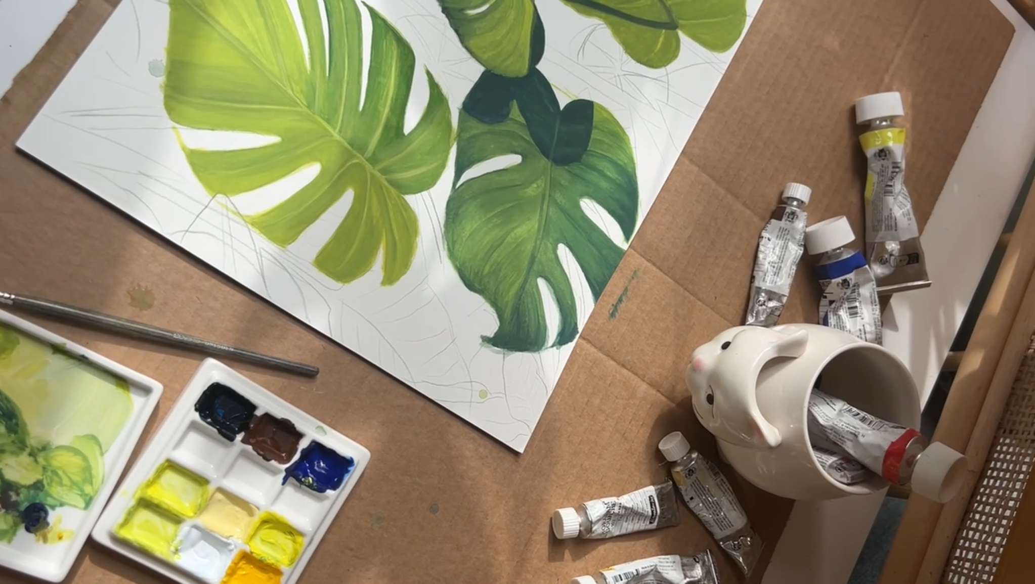 A partially completed painting of tropical green monstera leaves on paper, with watercolor paints, tubes of paint, a paint palette with yellow, green, and blue colors, and a ceramic mug shaped like a bunny nearby on a table.