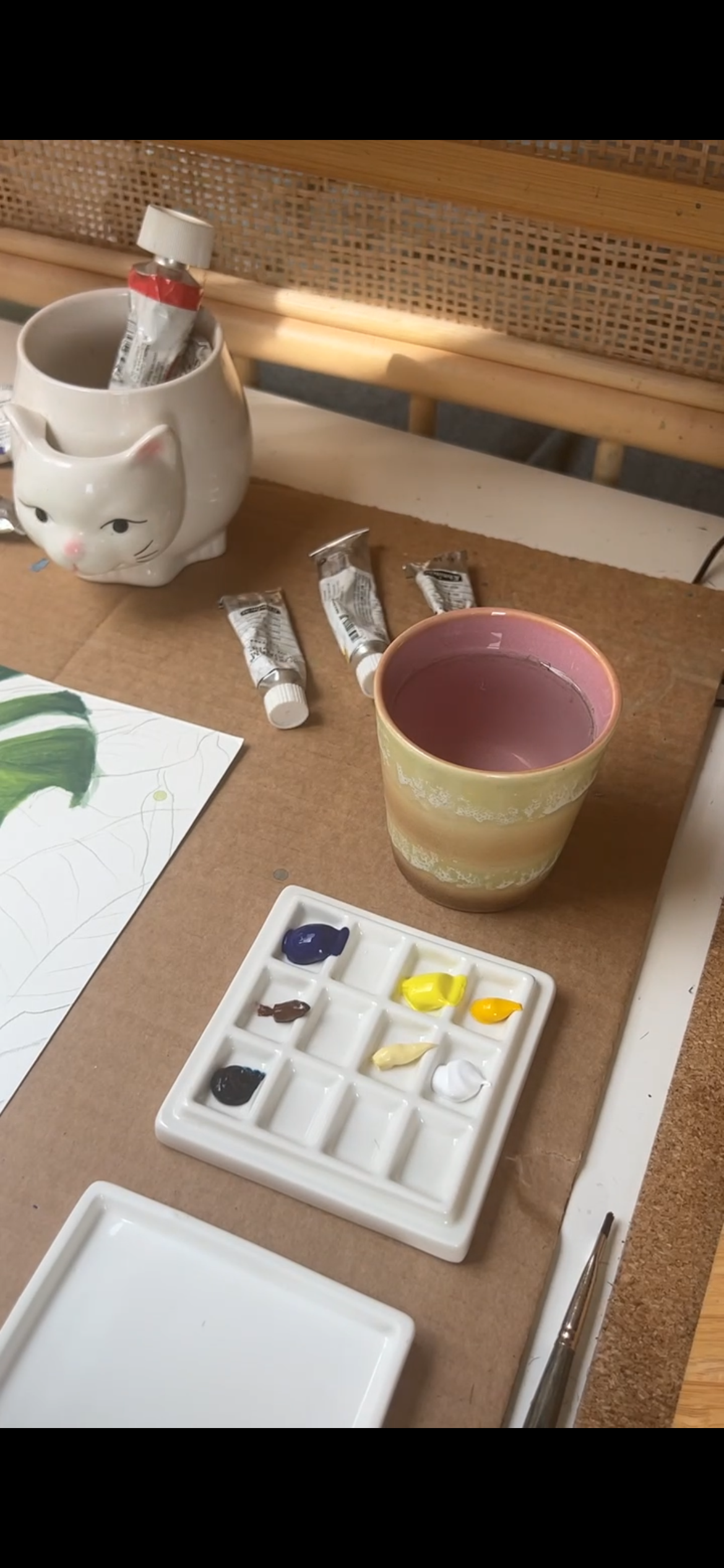 An art workspace with a paint palette containing various colors, a ceramic mug shaped like a cat holding a paint tube, a ceramic cup of water or paint, and a watercolor painting of green leaves on paper.