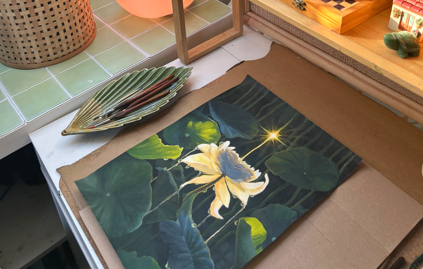 A painting of a white flower with green leaves, a star-shaped light, and dark background on a table with art supplies and decorative objects around.