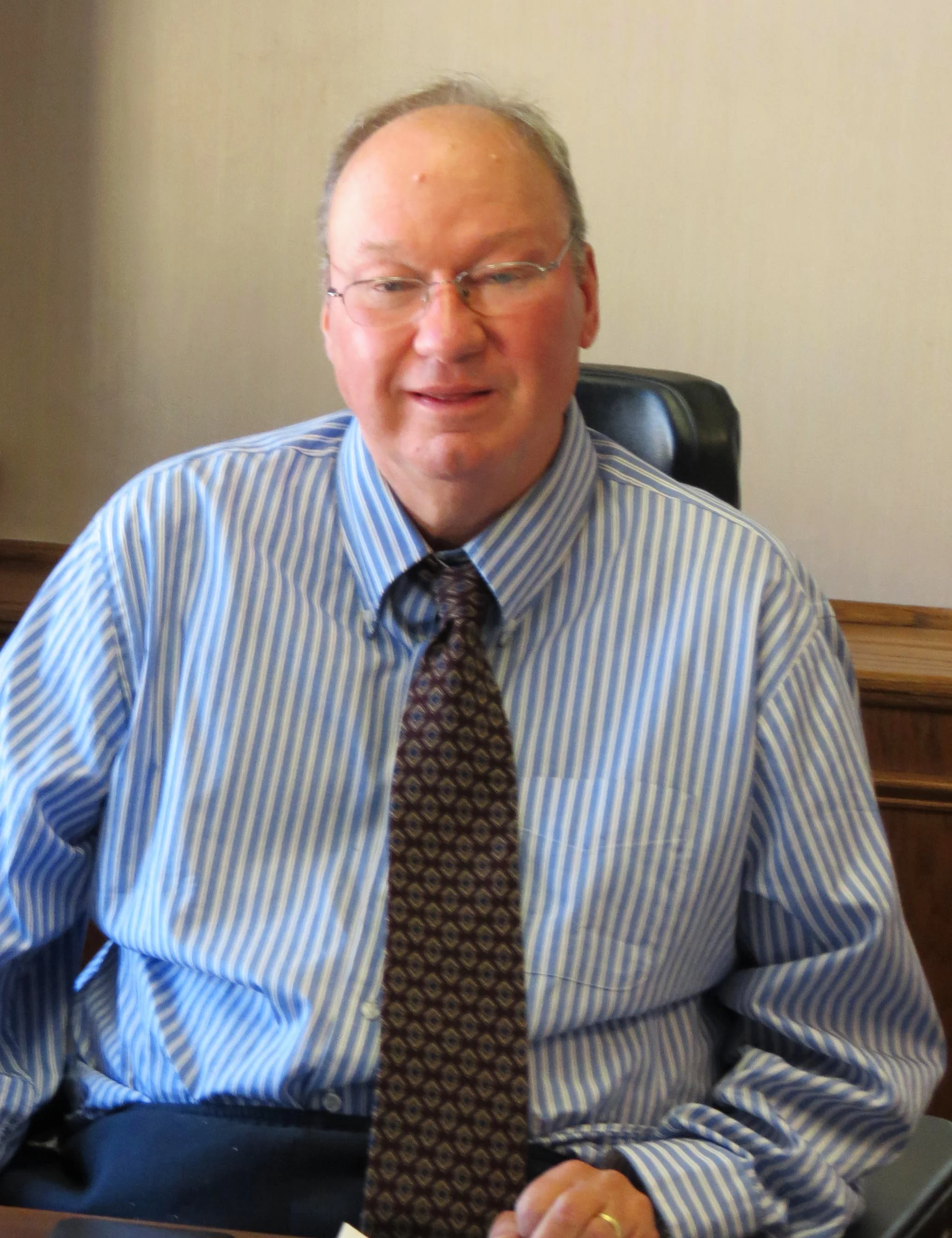 A middle-aged man with glasses, dressed in a blue striped shirt and tie, sitting at a desk in an office environment.