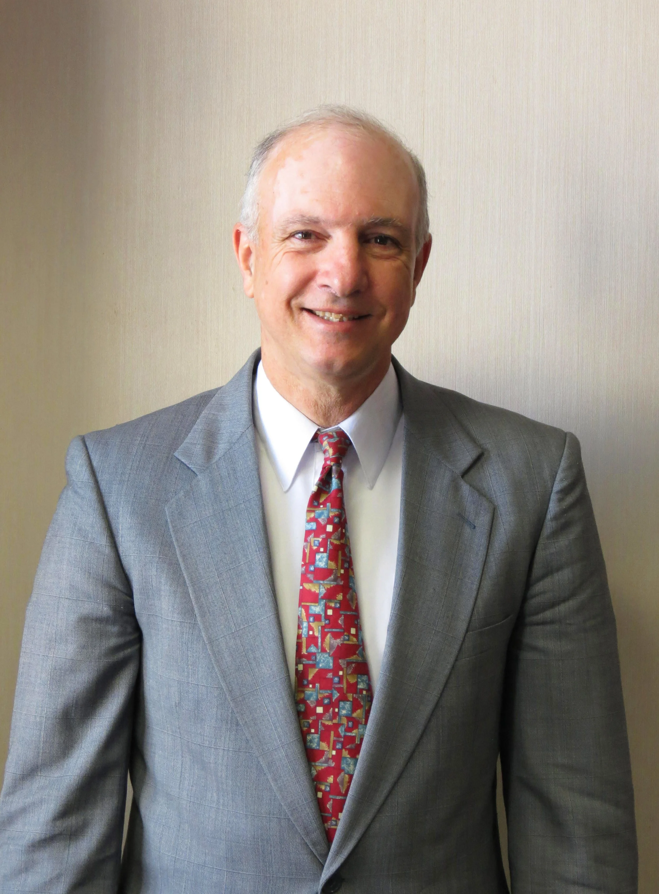 A middle-aged man wearing a gray suit, white shirt, and a colorful patterned tie, smiling and standing against a plain light-colored wall.