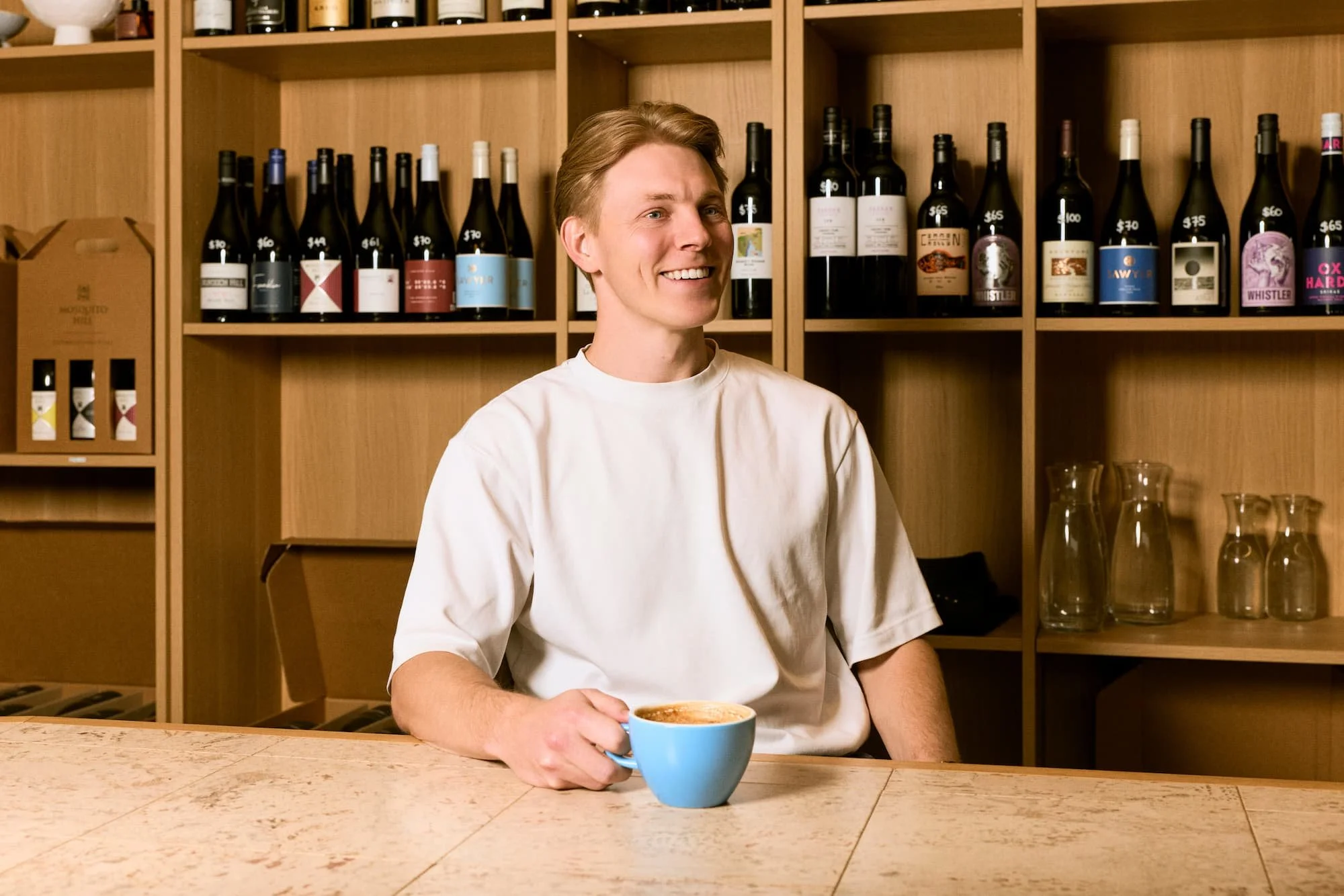 A smiling man sitting at a bar or counter holding a blue cup of coffee or tea. Behind him is a wooden shelf filled with bottles of wine and liquor, and to the right, clear glassware is visible.