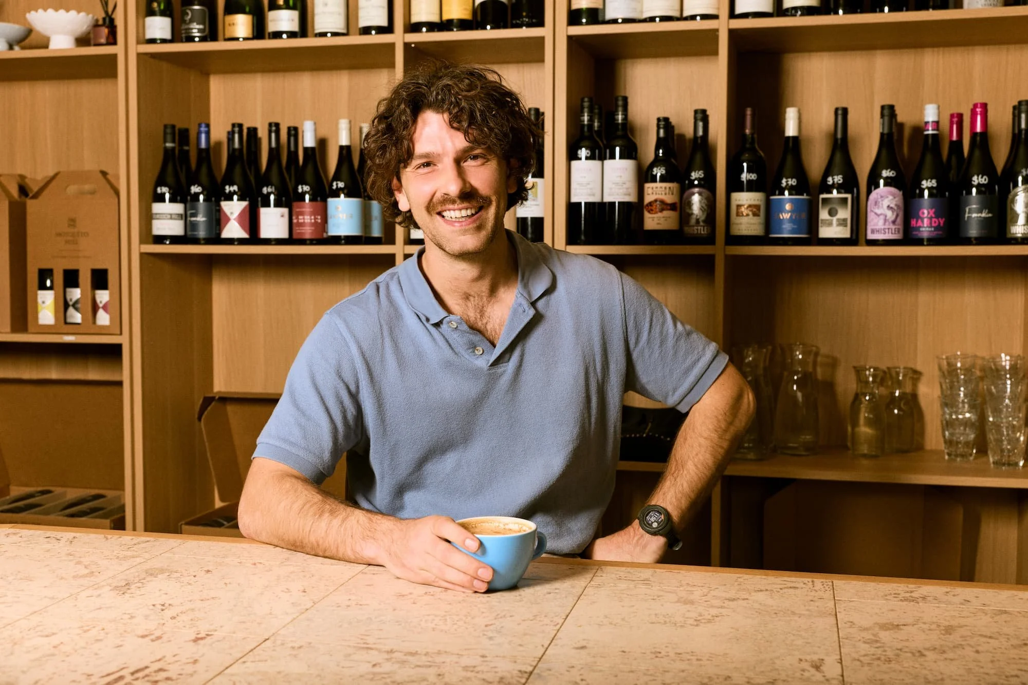 A man with curly hair and a mustache smiling while holding a blue coffee cup in a wine shop or bar with shelves of wine bottles and glassware in the background.