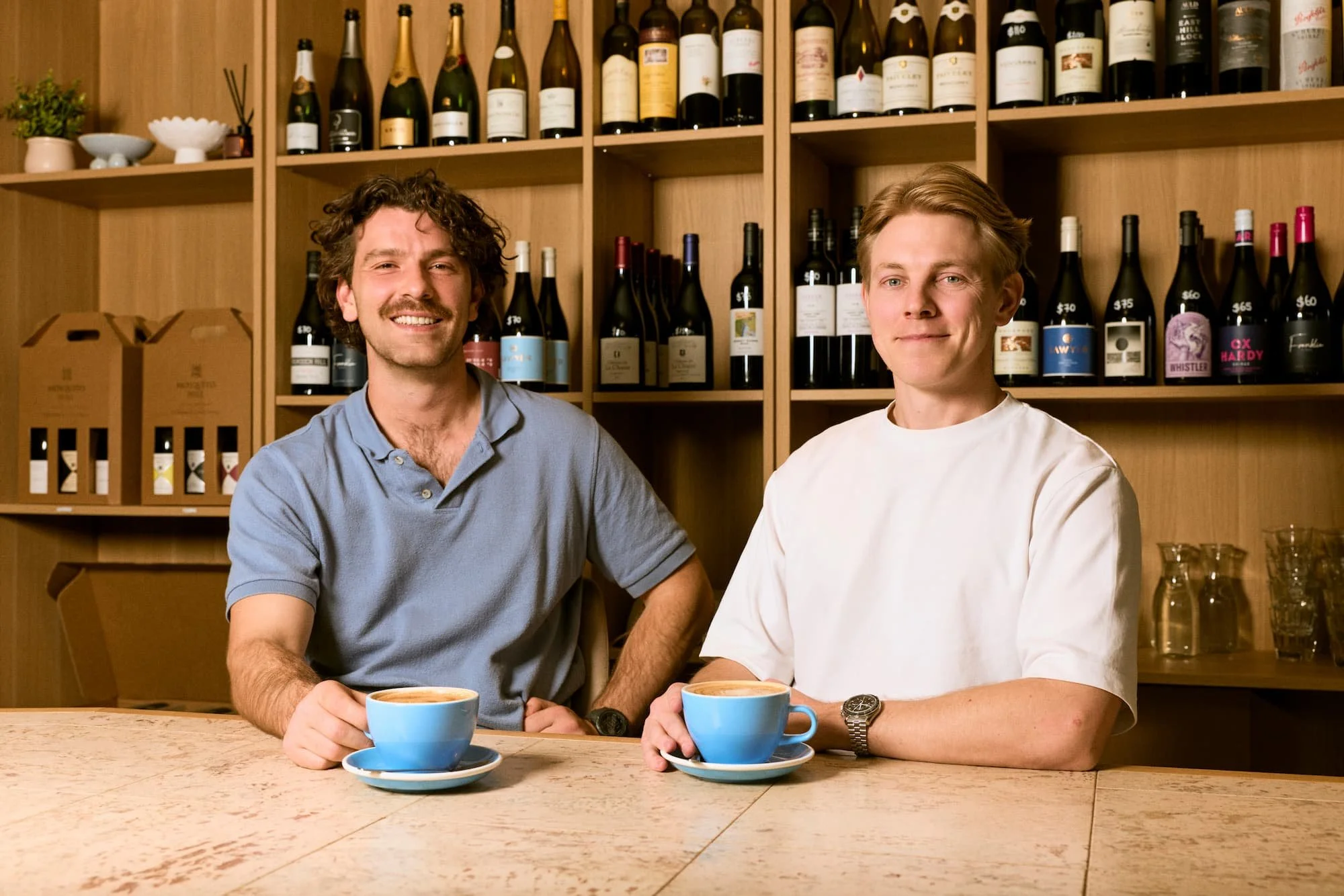 Two men sitting at a table with blue coffee cups, in a wine cellar or bar with wooden shelves filled with wine bottles behind them.