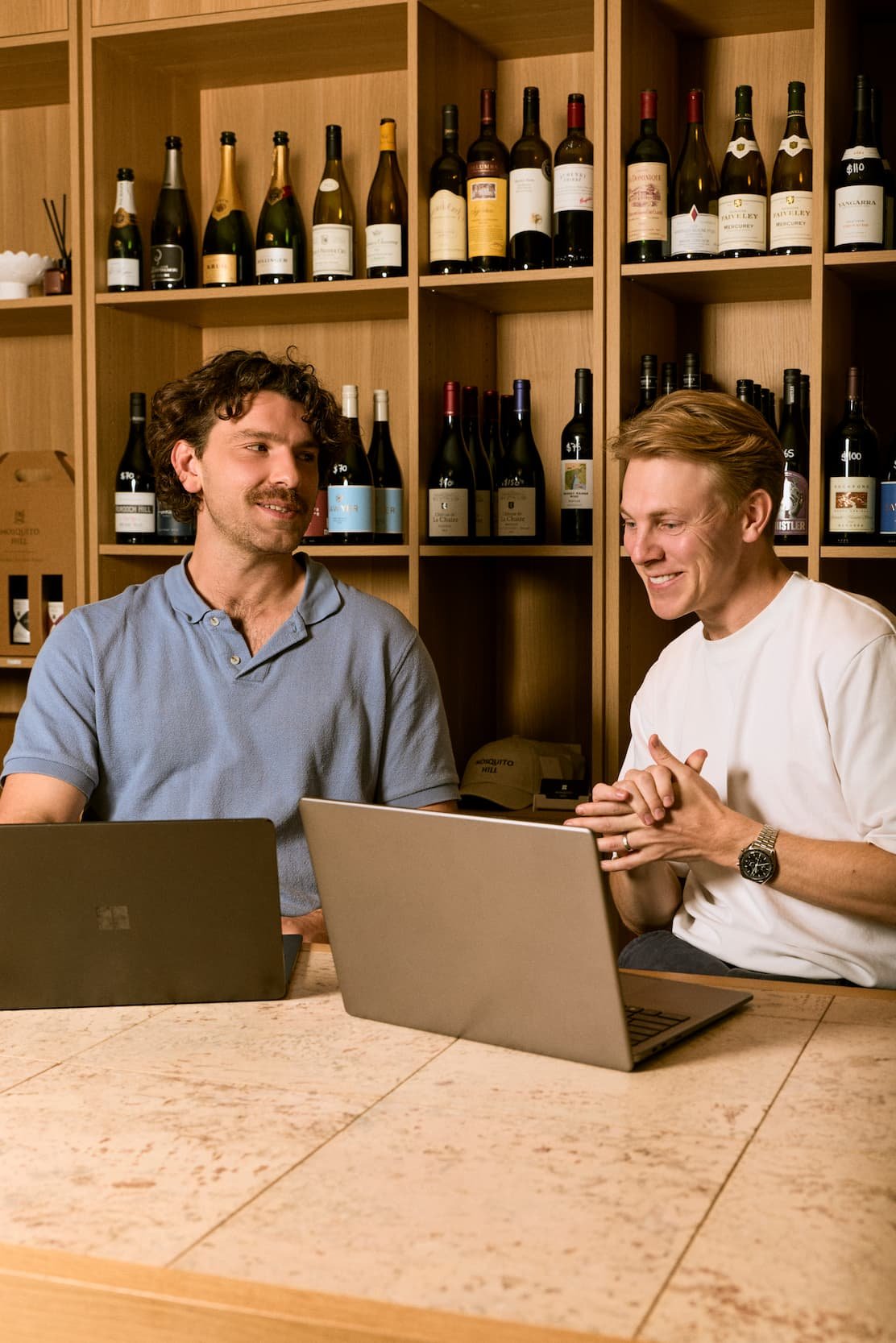 Two men sitting at a table with laptops, talking and smiling, with wine bottles on shelves in the background.