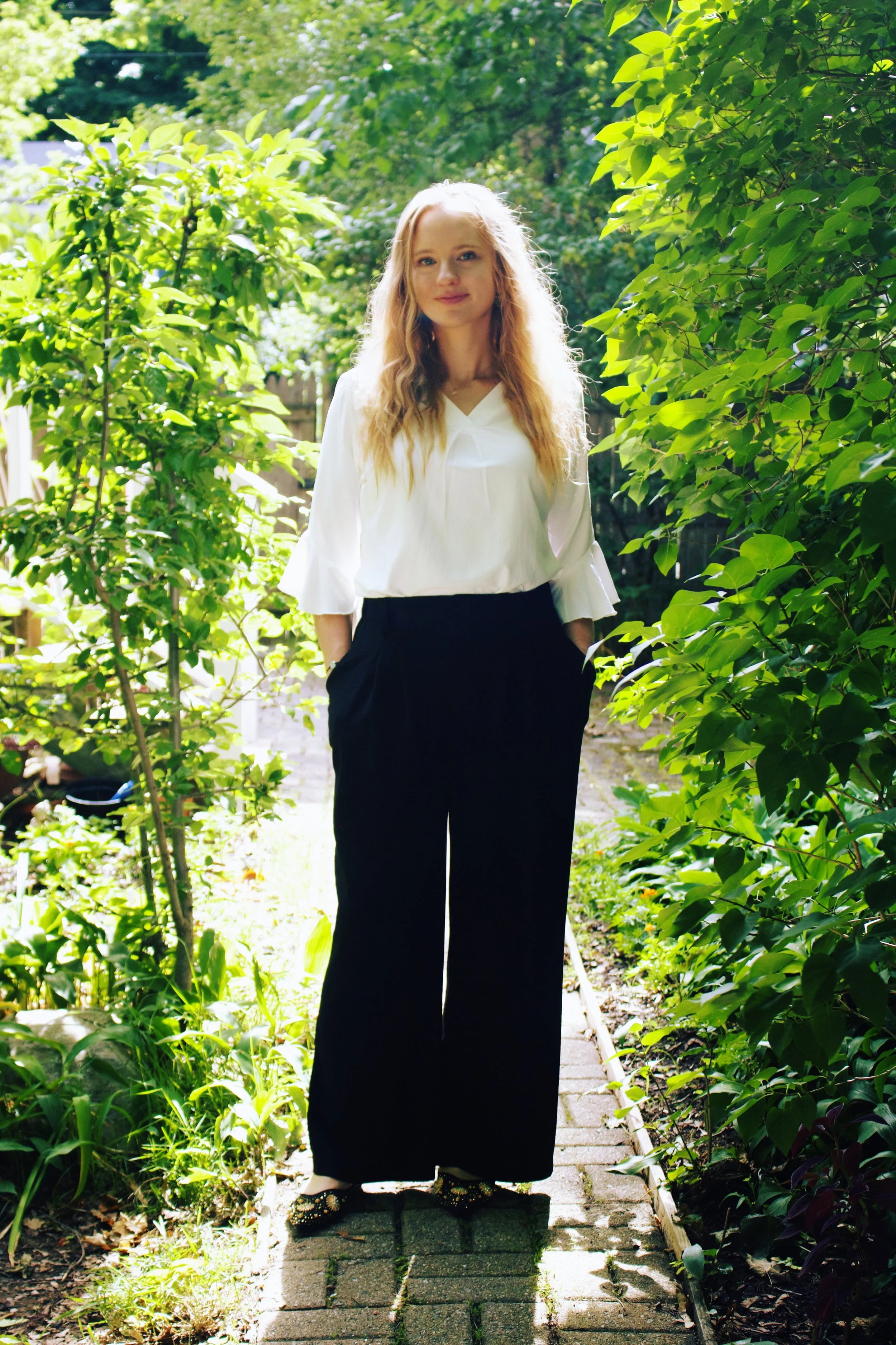 Young death doula with long blonde hair, dressed in a white blouse and black wide-leg pants, standing on a small brick ledge in a lush, green garden with sunlight filtering through the trees.