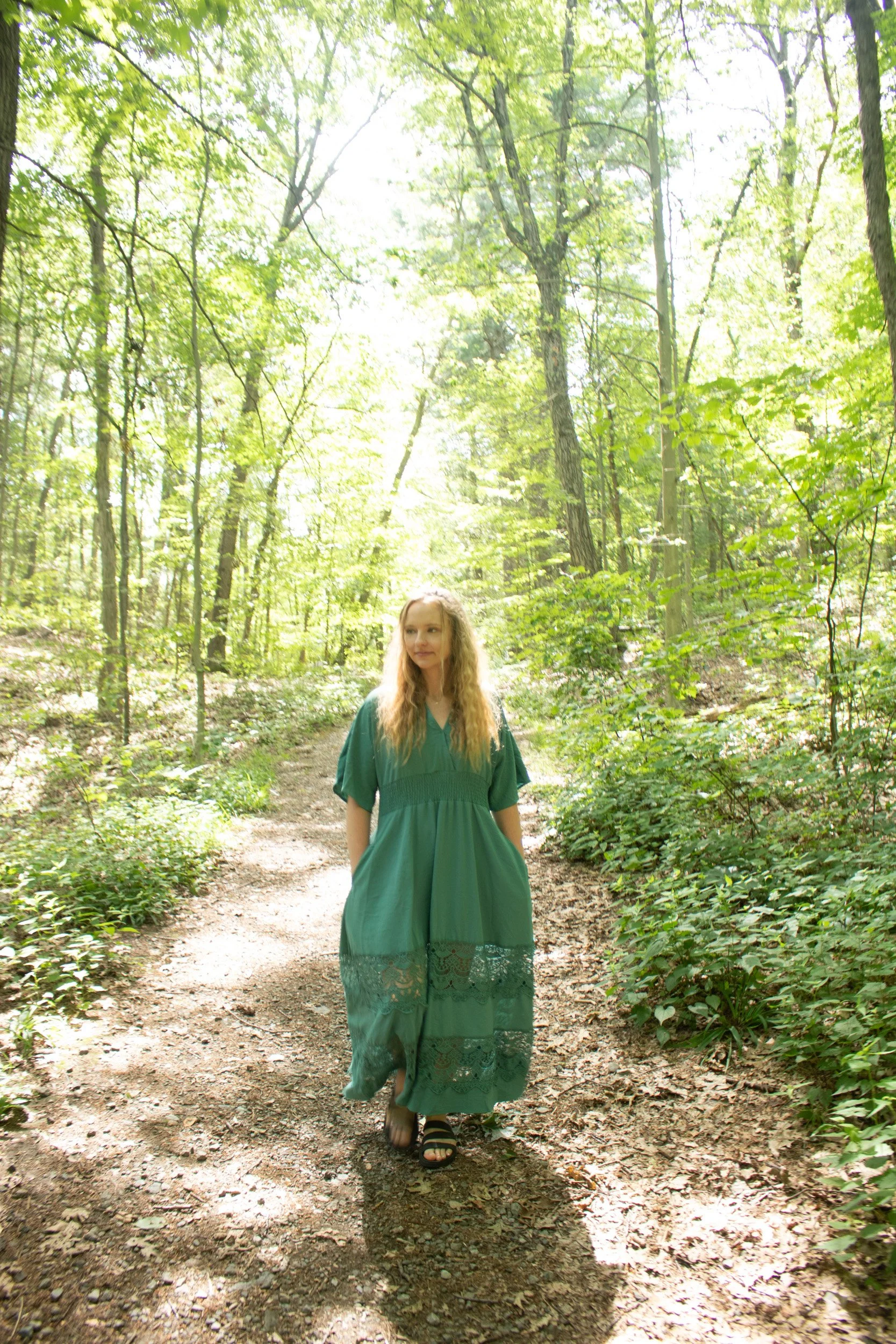 A young woman, a death doula, with long blonde hair walking on a forest trail, wearing a green dress with lace details at the hem and black sandals in Massachusetts.