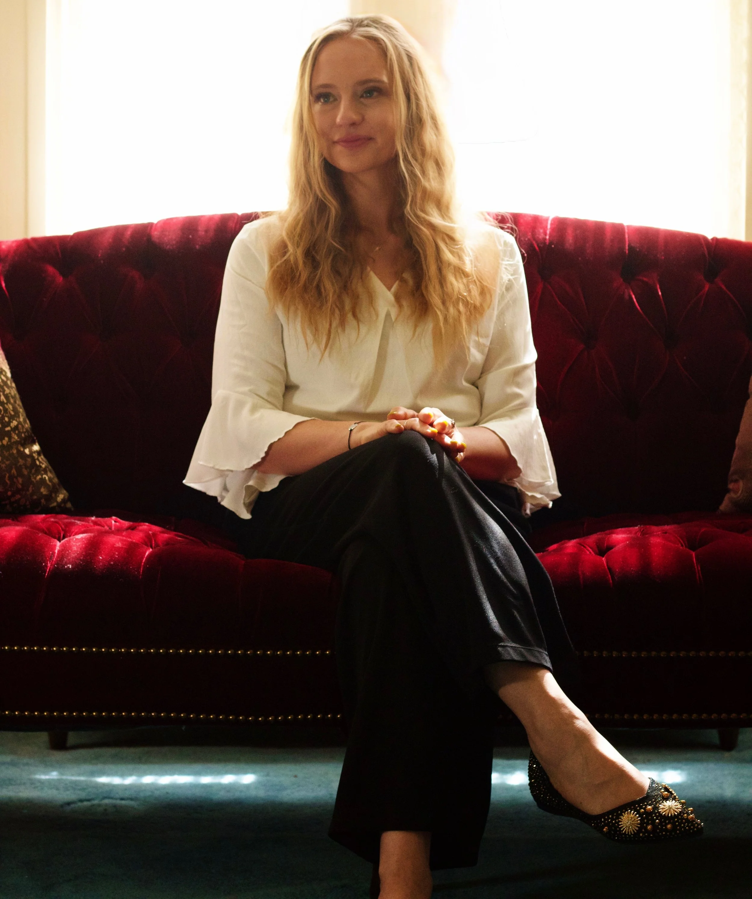 Woman, a death doula, sitting on a red velvet sofa with a bright backlight behind her, wearing a white top and black pants, with gold and black embellished shoes.